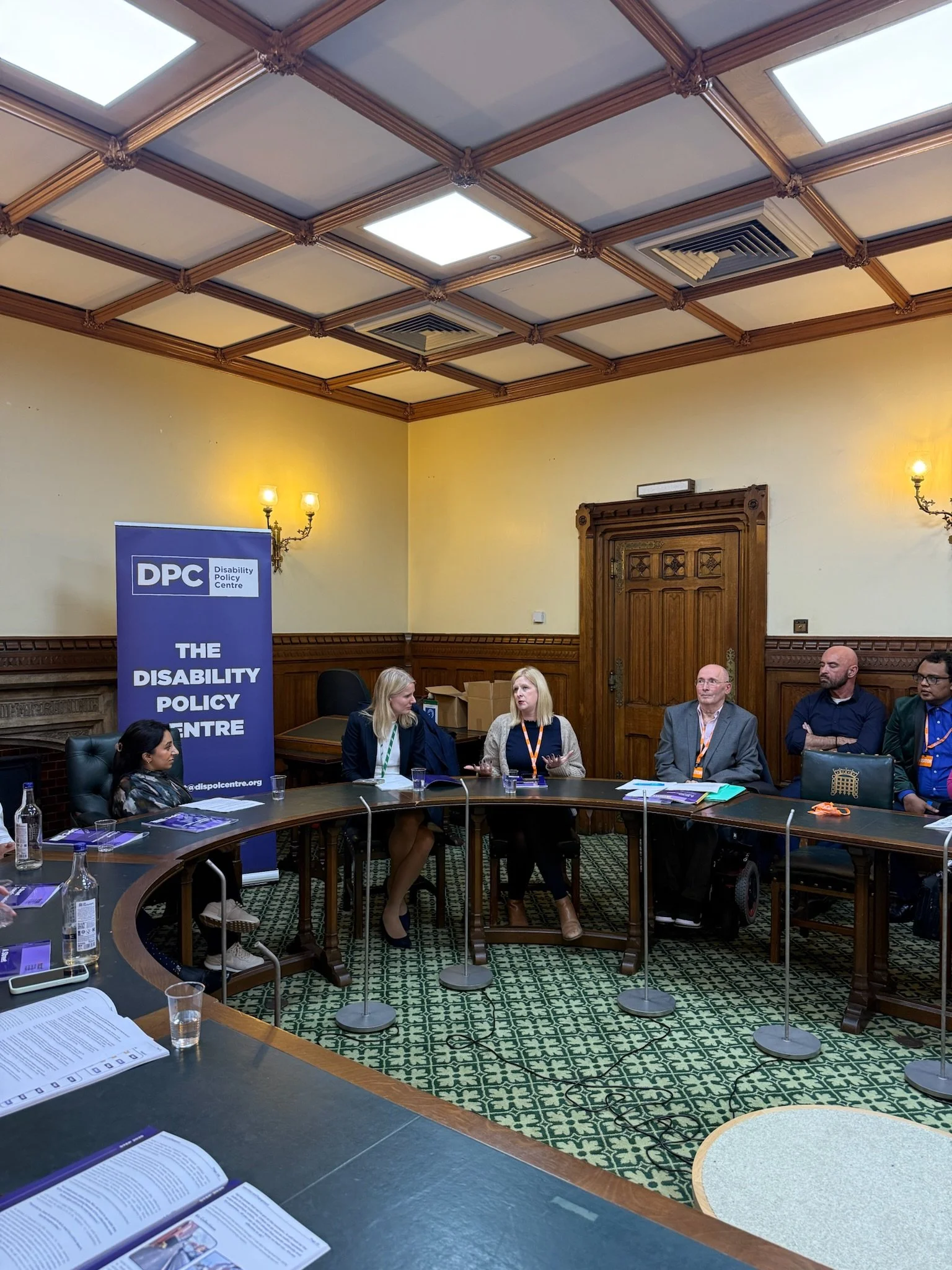 Panel discussion in a parliamentary room hosted by the Disability Policy Centre. At the front, from left to right: Dr Shani Dhanda (chair), Rebecca Paul MP, Louise Connop, and Ian Oakley, seated beside a DPC banner.