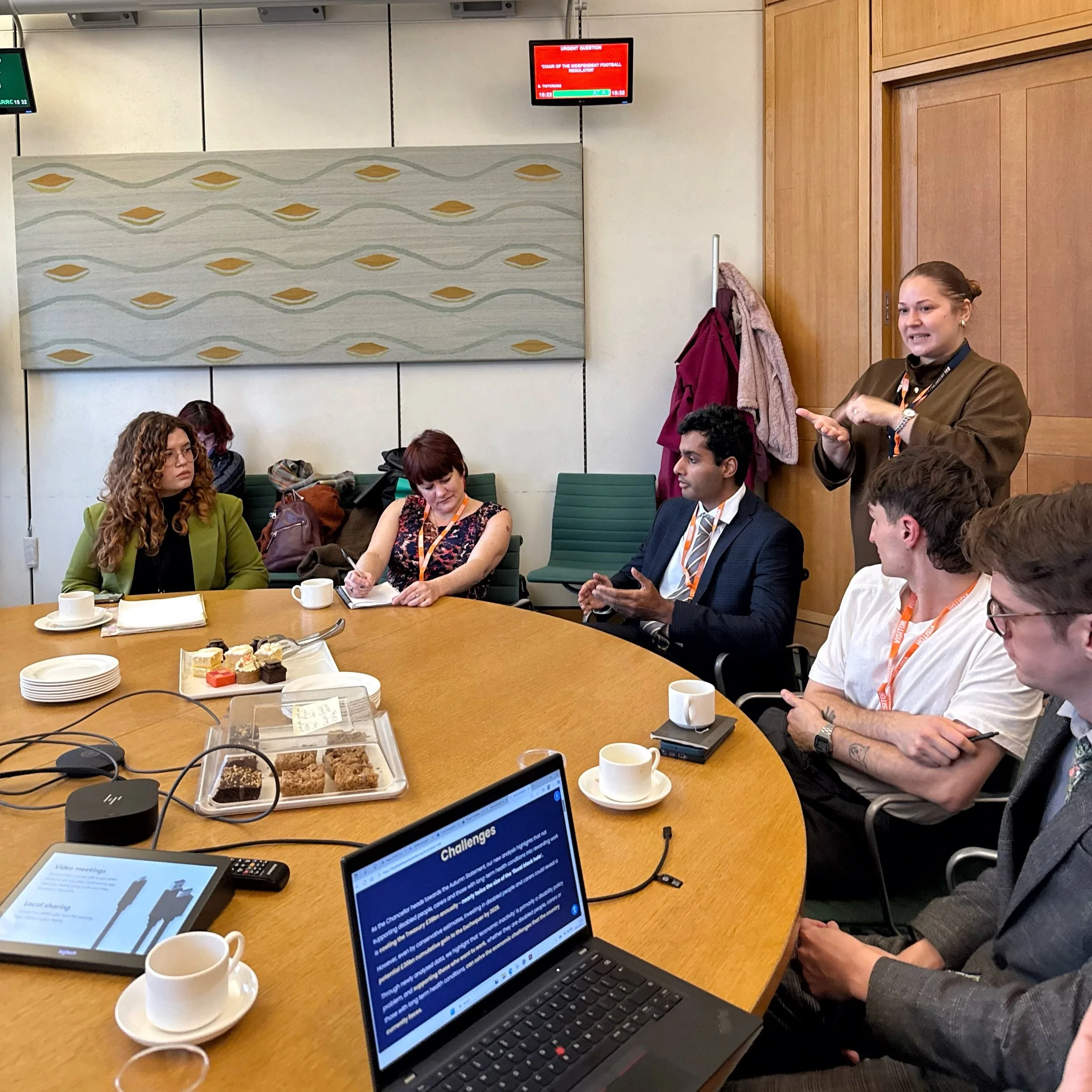 A roundtable meeting in Parliament. From left to right: Bethany, Ellen, Arun and Mikey engaged in discussion, with a BSL interpreter standing behind them. Cups, notes and devices are on the table.