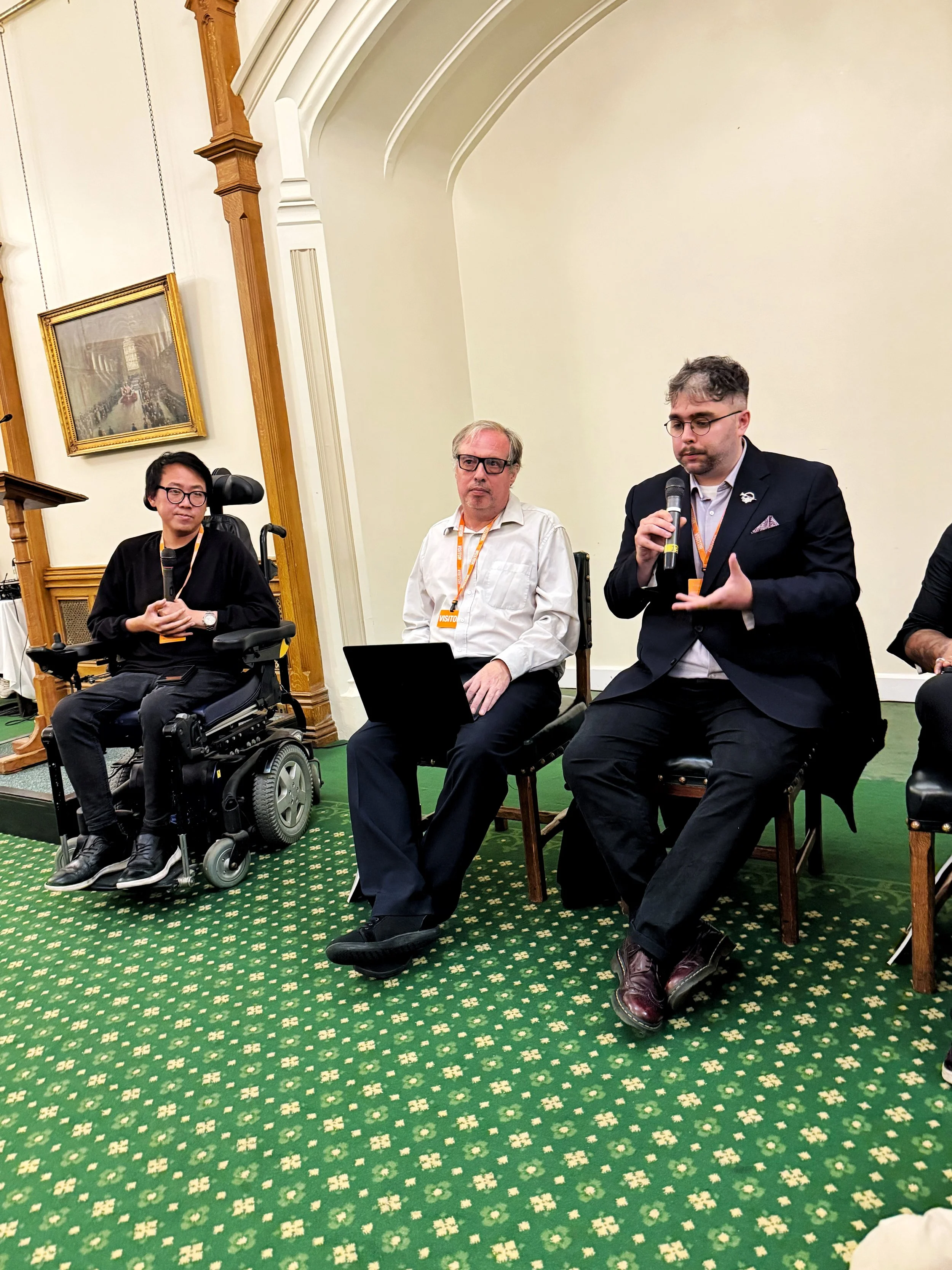 Panel discussion in the Churchill Room, Houses of Parliament, with James seated in a wheelchair and Gáibhin McGranaghan speaking into a microphone, and Mike sat in the middle of them with a laptop on his lap.