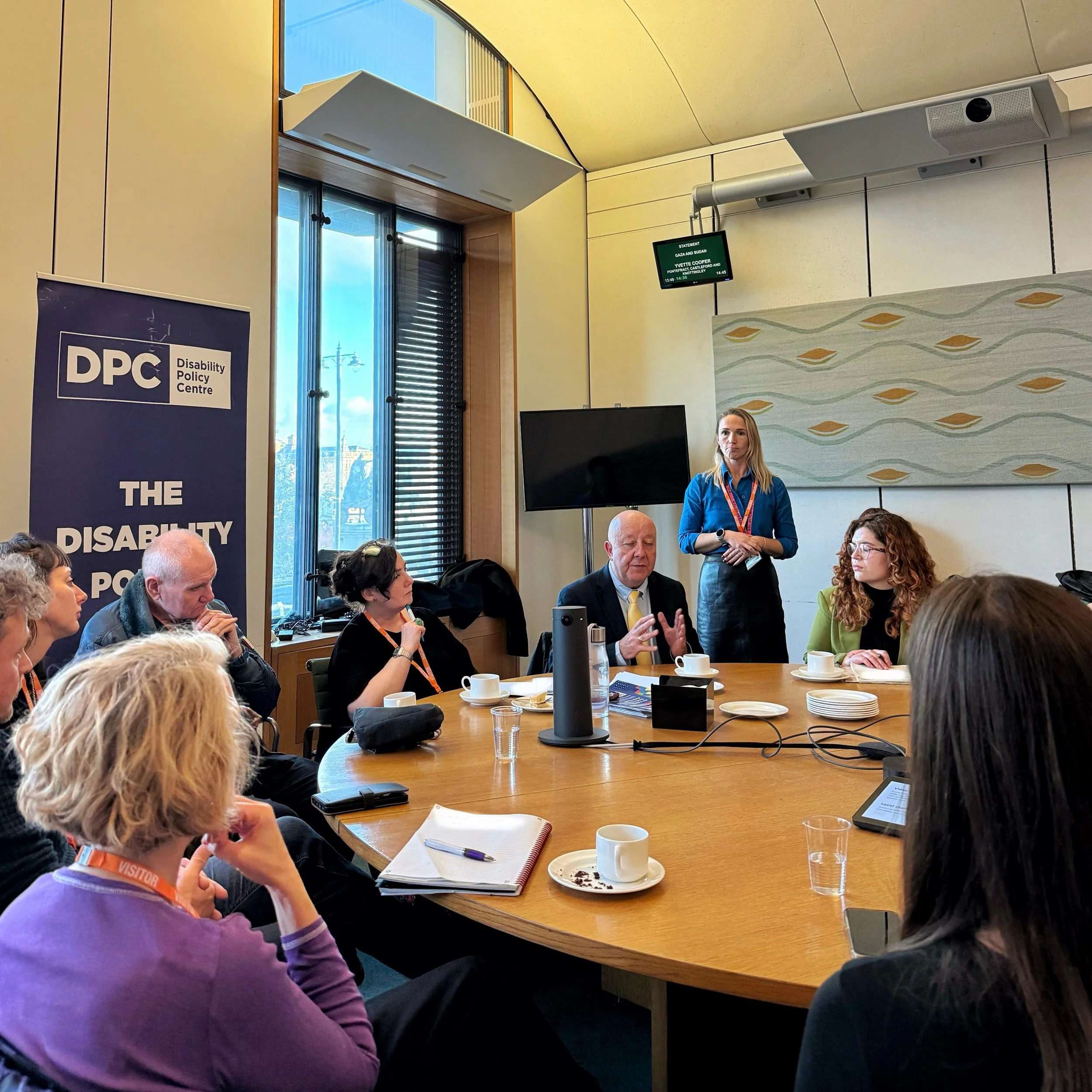 A roundtable meeting in Parliament. Steve speaks at the table, with Maria to his left and Bethany to his right. A BSL interpreter stands behind him, alongside the DPC banner and discussion materials.