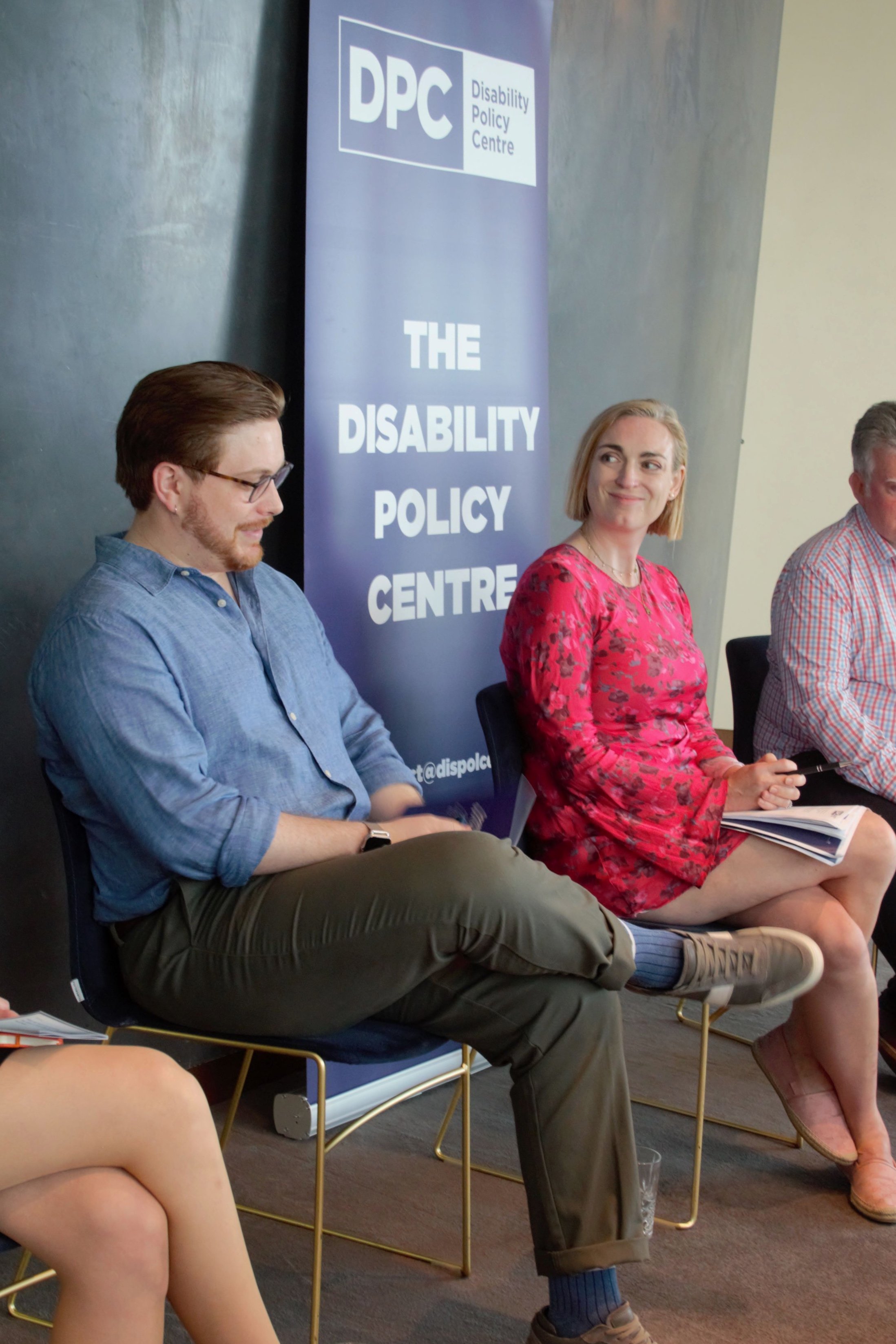 Photograph of Joseph and Alys sat down on the panel. They are sat infront of a DPC purple banner. Alic is on the right in a pink dress looking towards Joseph who is dressed in a blue shirt and khaki trousers.