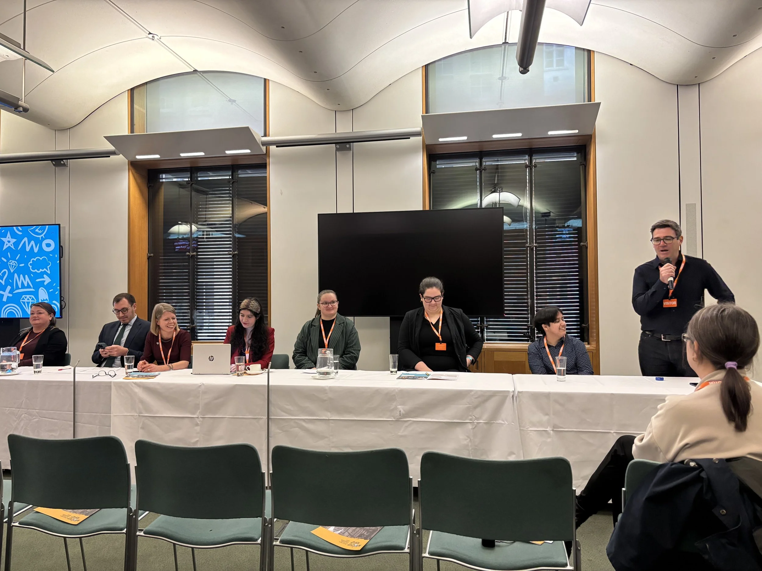 A photograph of a panel discussion in parliament. At the far right Andy Burnham is stood up talking into a microphone. He is wearing a black shirt and trousers.