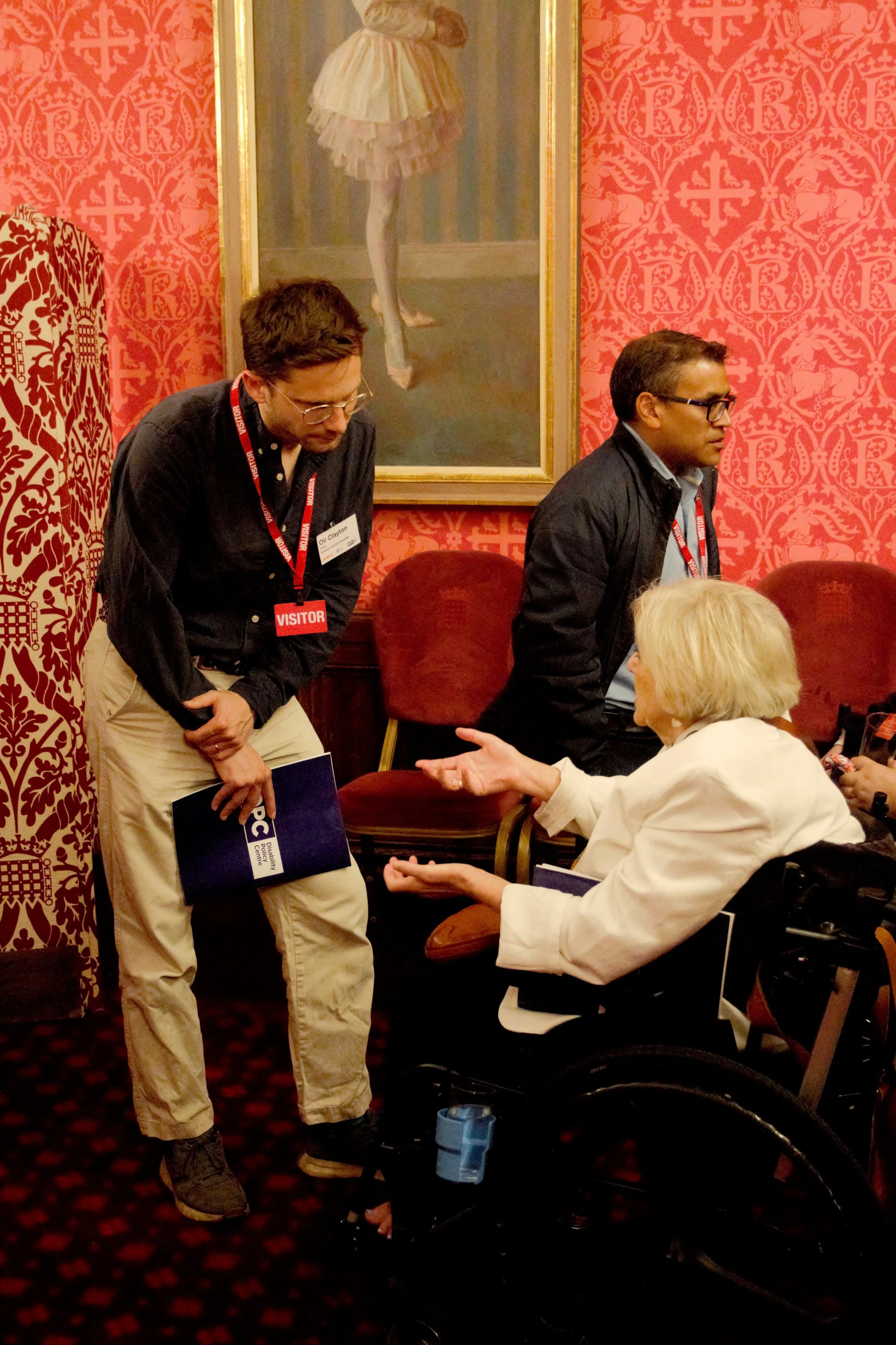 Two guests conversating, woman on the right in a wheel chair, with blonde hair in a white suit jacket, man on the left in beige trousers, navy buttoned up shirt and red visitors langyard leaning down to speak the woman. There is a portrait of a balle