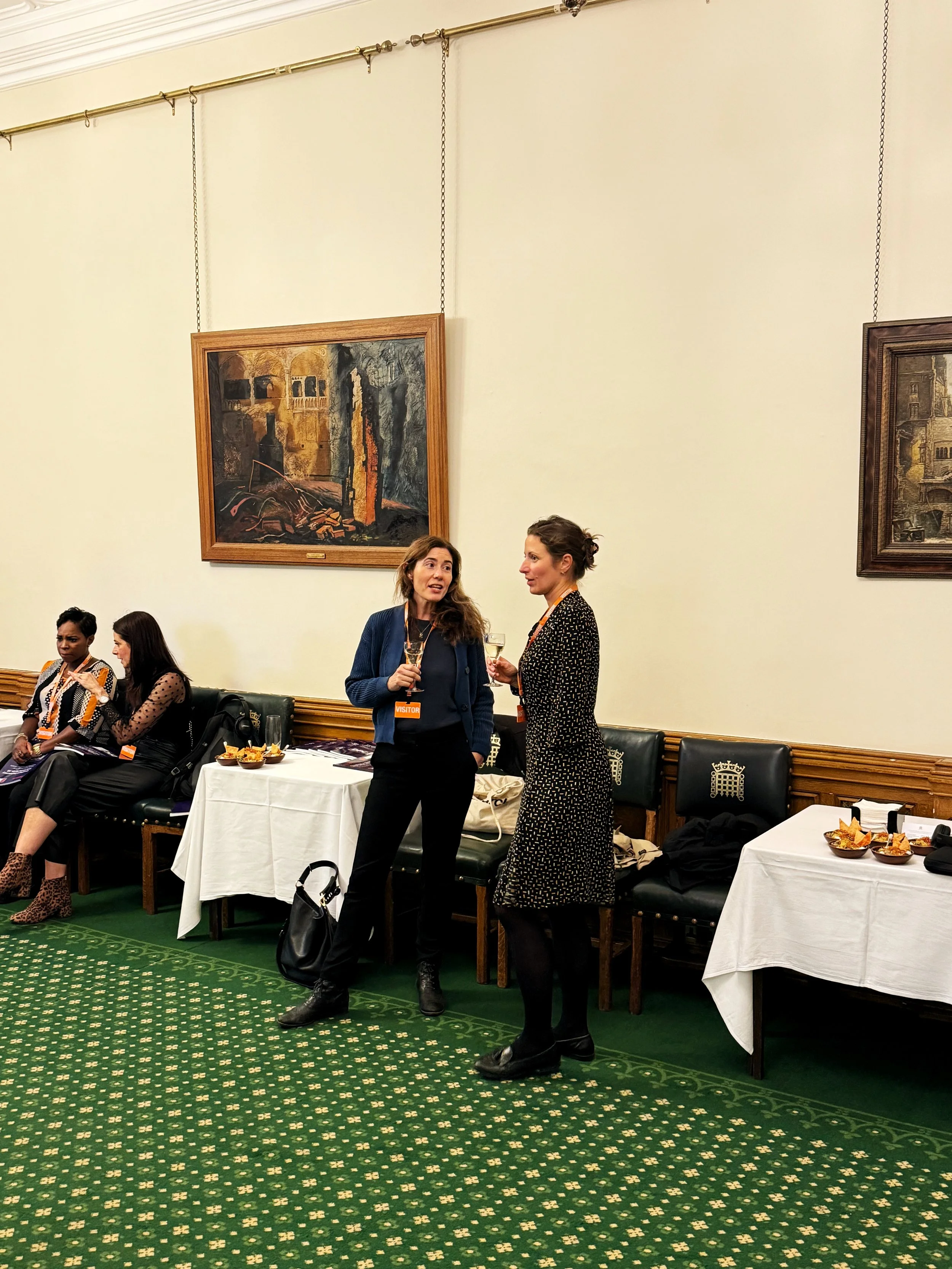 Two guests talking in the Churchill Room, Houses of Parliament, holding drinks in front of wall paintings. Other attendees are seated nearby, with tables set with refreshments and green patterned carpet beneath