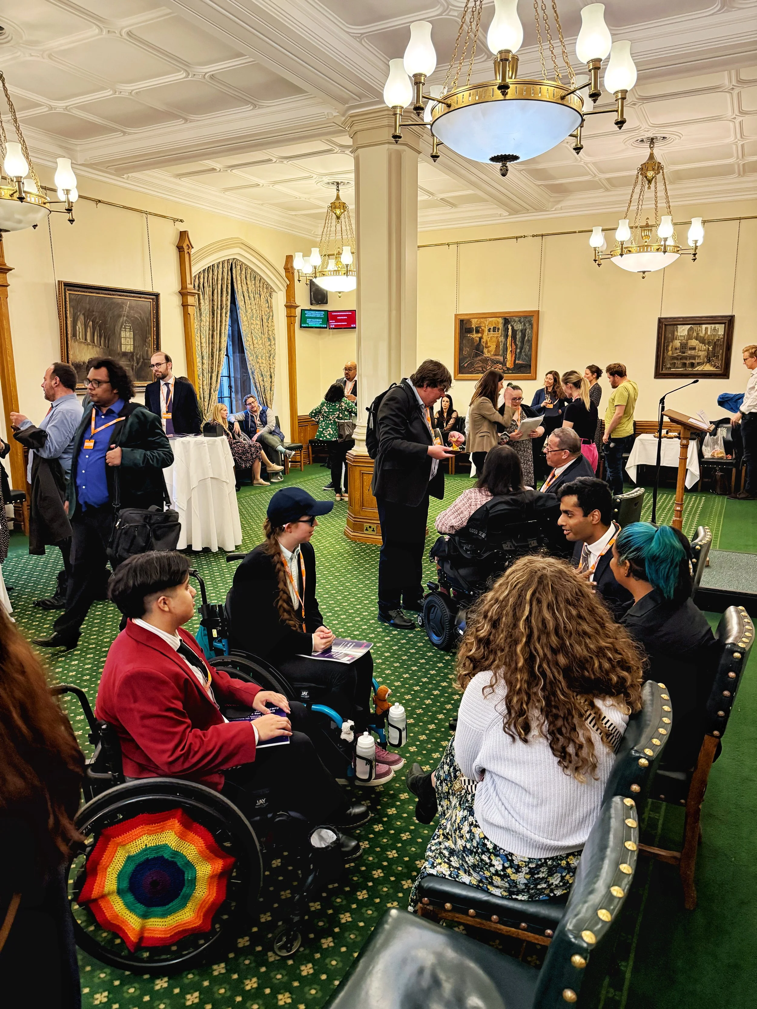 Busy scene in the Churchill Room, Houses of Parliament, with groups of people networking and talking. Several wheelchair users and seated guests are present, surrounded by chandeliers, paintings, and green patterned carpet.