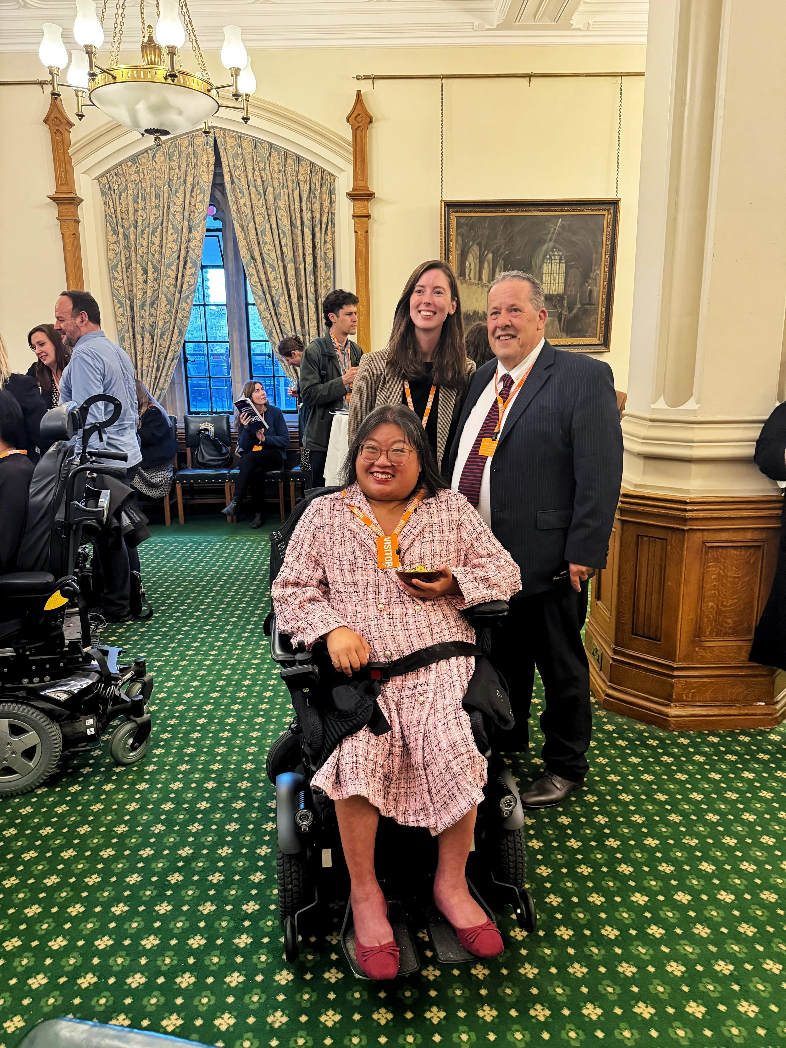 Three guests smiling for a photo in the Churchill Room, Houses of Parliament, including a wheelchair user in front. Other attendees mingle in the background beneath chandeliers, paintings, and tall curtains