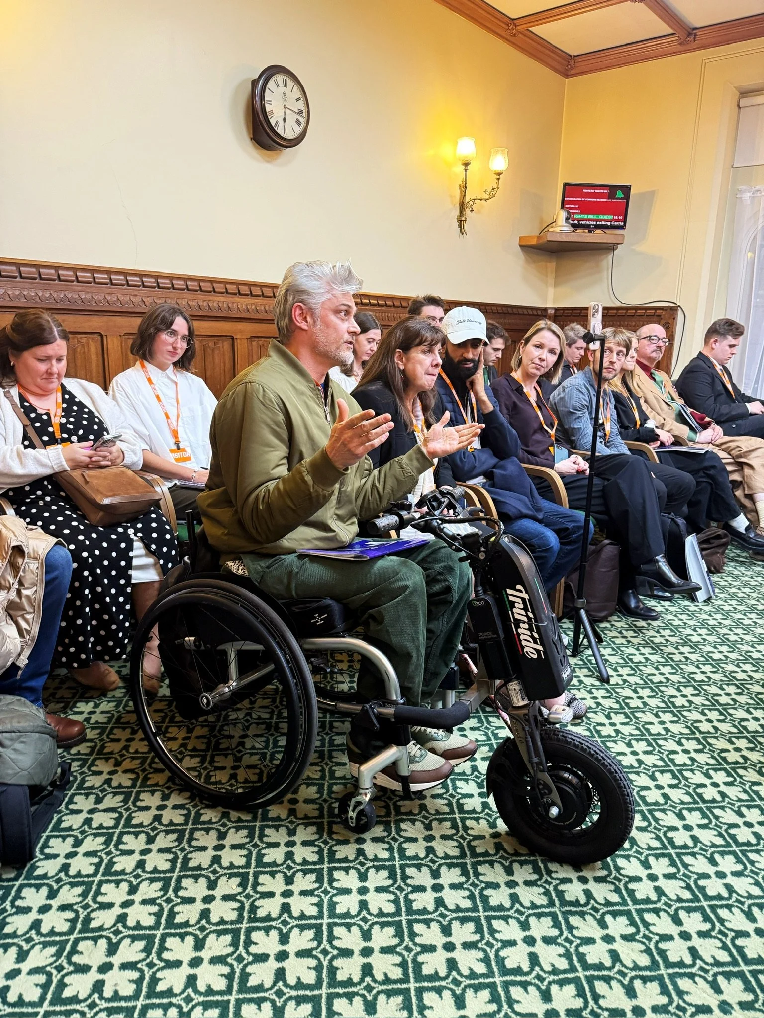An audience member using a powered wheelchair speaks and gestures during the discussion, surrounded by attendees seated and listening attentively in the parliamentary room.