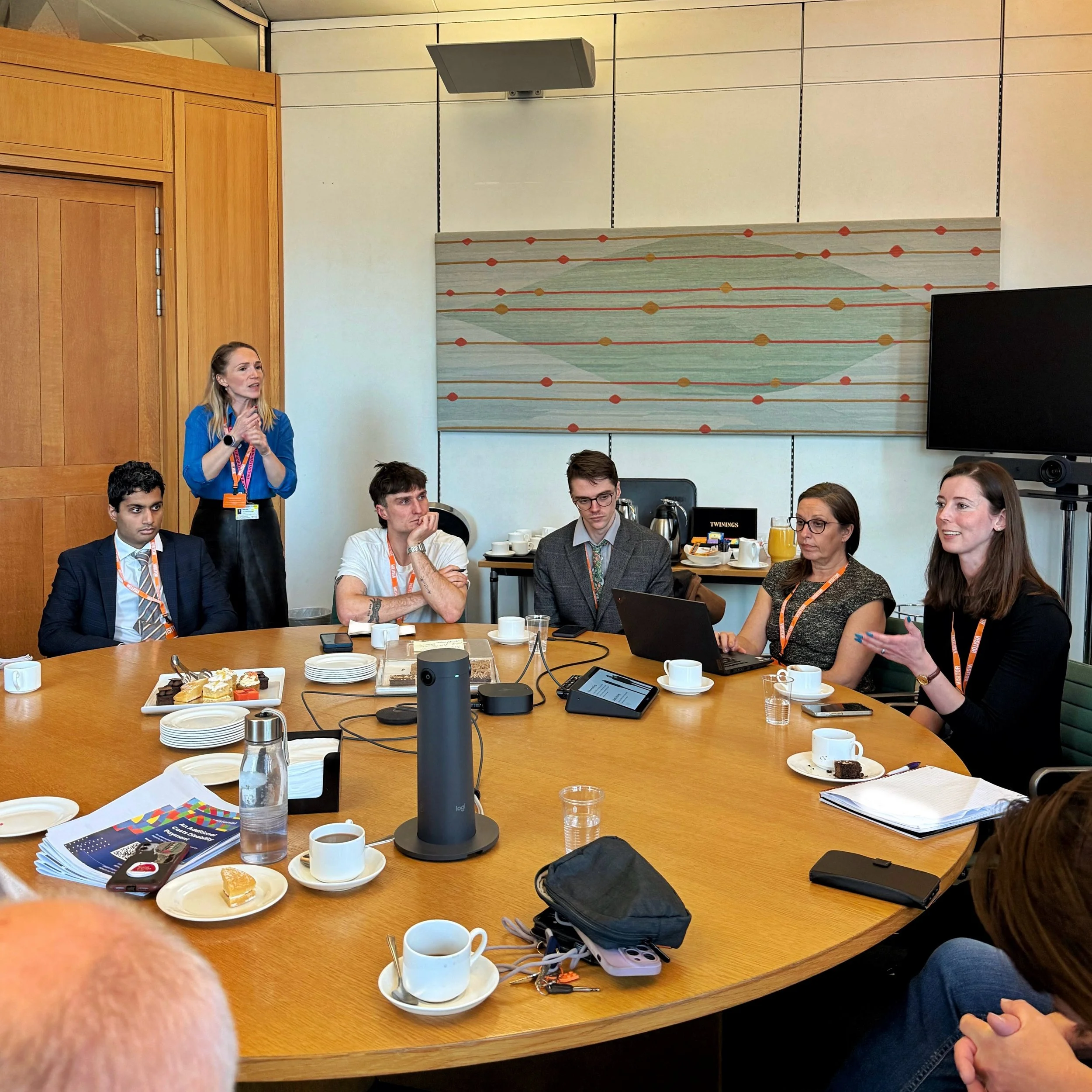 A roundtable meeting in Parliament. Chloe is speaking on the right, with Cara, Josh, Mikey and Arun seated to her left. A BSL interpreter stands behind, with notes and refreshments on the table.