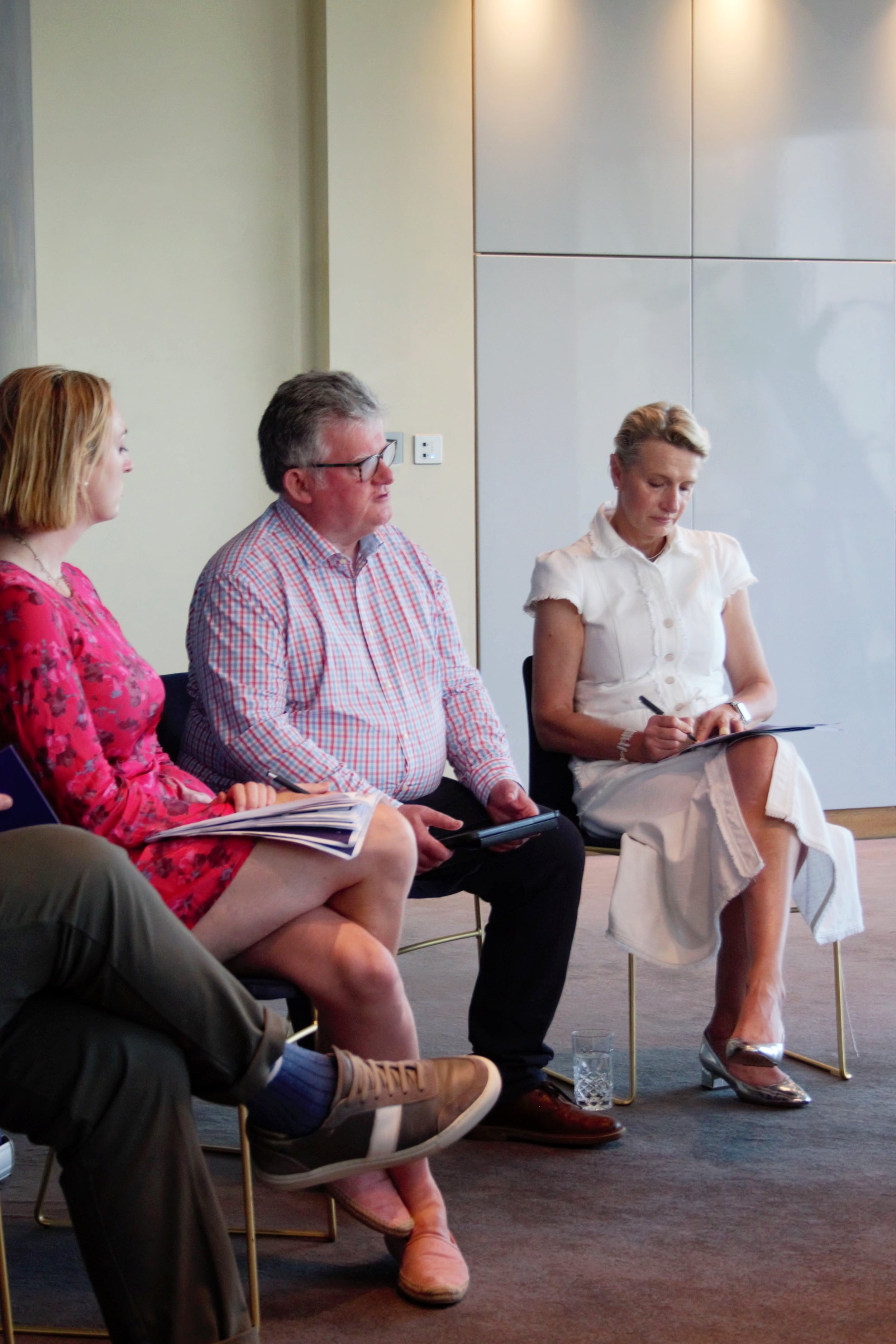 Photograph of Alys, Johnny and Alison sat down in row next to each in a panel format. Johhny is engaging with the audience. Alys is wearing a pink floral dress. Johhny is dressed in baby pick checkered shirt and black trousers. Alison is in a white l