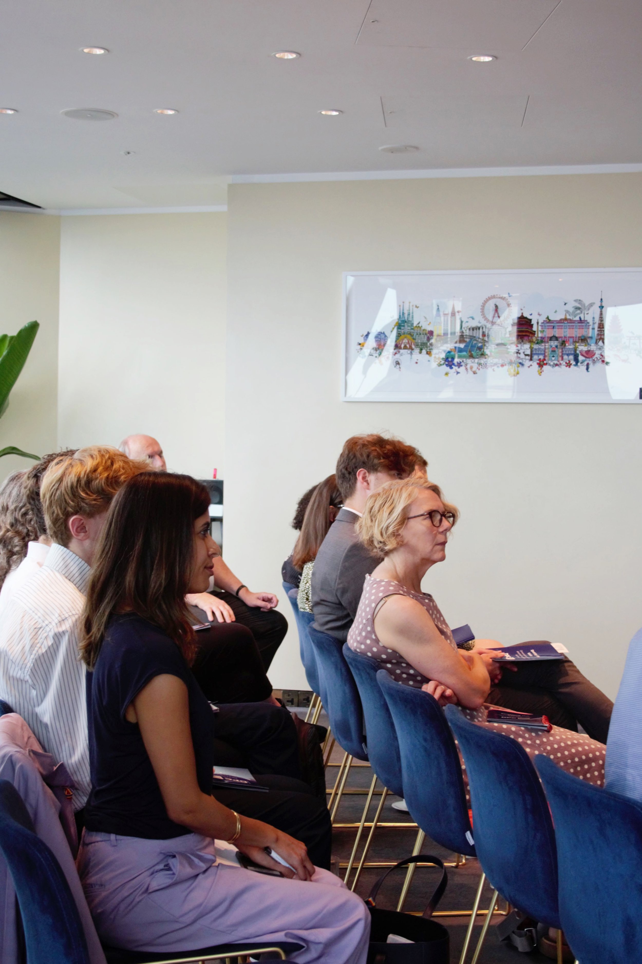 Photograph of audience seated in two rows on blue chairs listening to the panel.