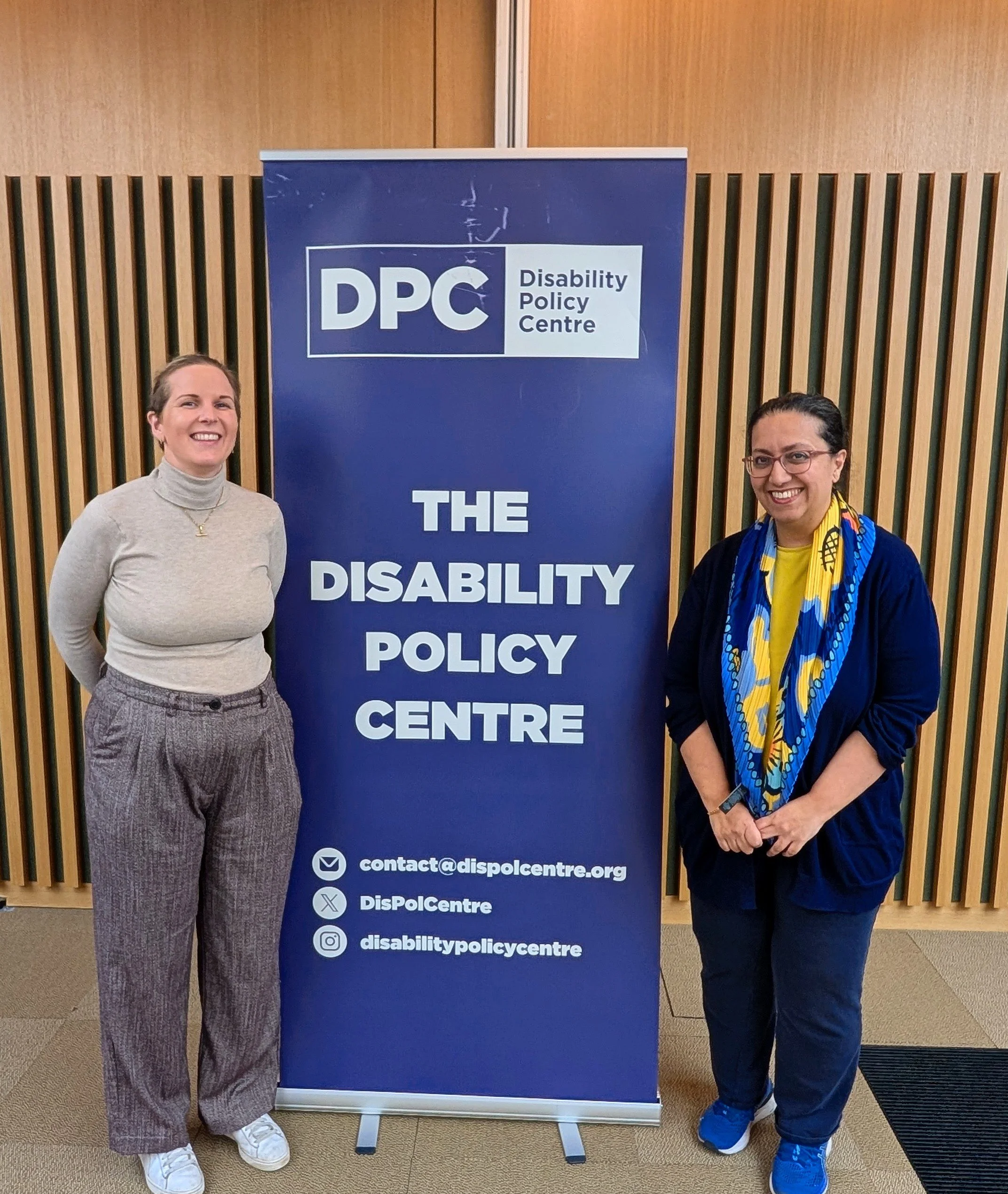 Emma stands beside Hina at City Hall, smiling for a photo. She has blonde hair and wears a beige jumper. Hina wears blue. They stand next to a Disability Policy Centre banner.