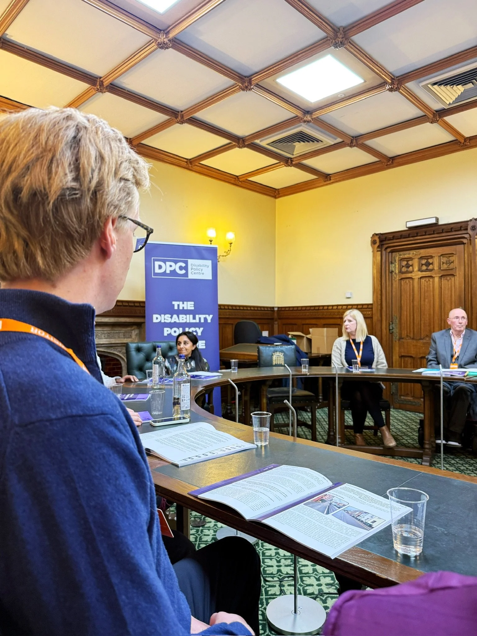 An attendee reading the A Street Transformation report during the event, with Dr Shani Dhanda, Louise, and Ian visible at the front beside a Disability Policy Centre banner.
