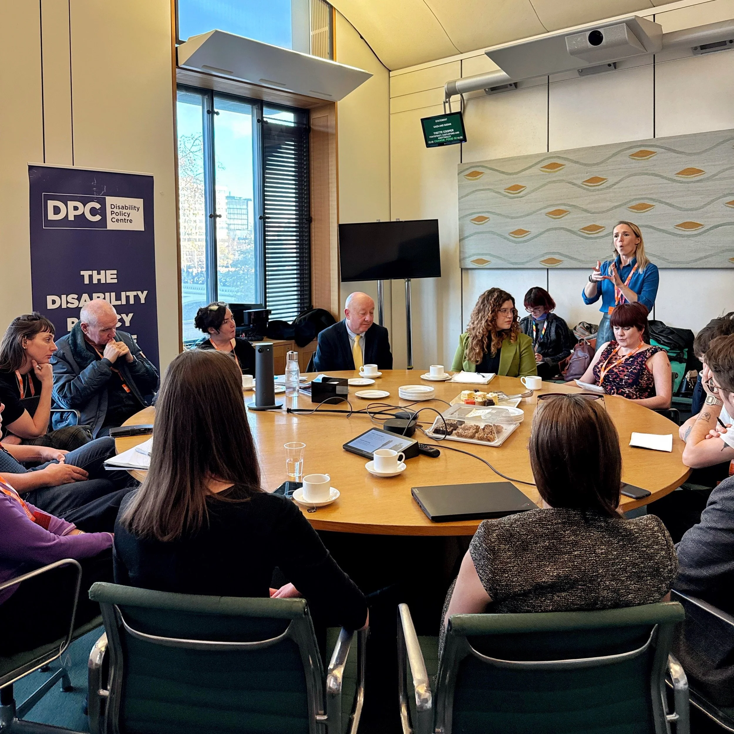 A roundtable meeting in Parliament. Ellen is speaking at the back right, with Bethany to her left, then Steve and Maria beside her. Other guests sit around the table with notes and refreshments.