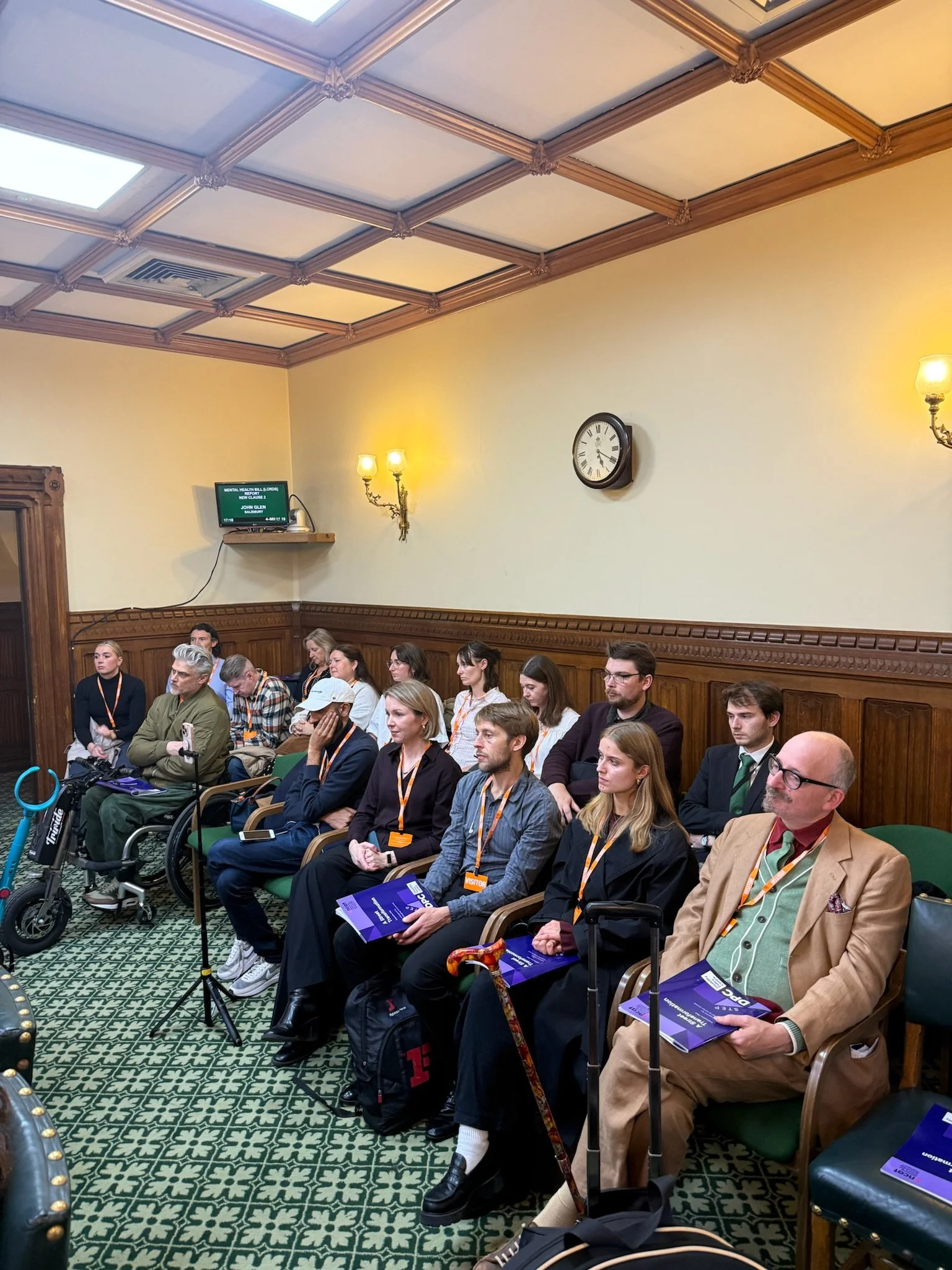 Photograph of 2 rows of audience members seated at the back of a Parliament room, all looking towards the front of the room. There are reports on peoples laps and a clock and lights on the wall behind.