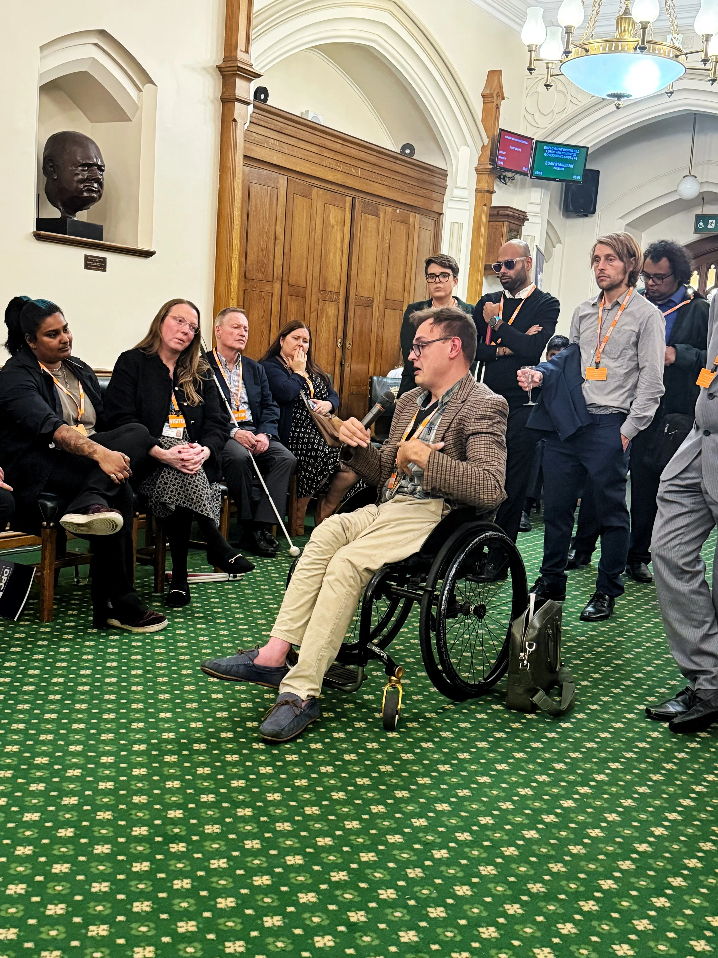 A male guest in a wheelchair speaks into a microphone during discussion in the Churchill Room, Houses of Parliament, while panelists including Ria Patel and Michelle Anglesea listen, with attendees standing nearby.