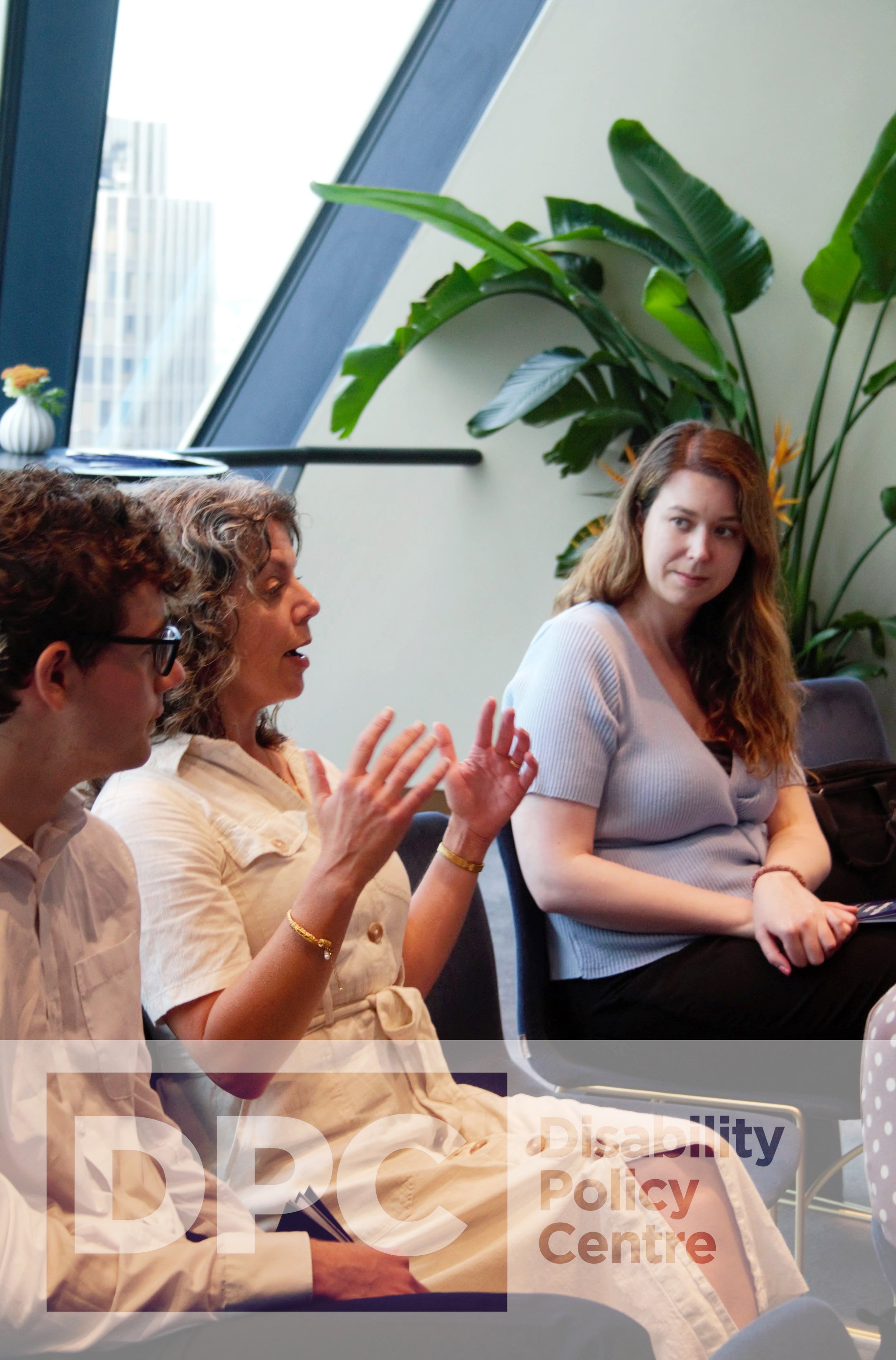 Close up photograph of a woman speaking from the audience. She is sat in the middle of two other guests, a female to the right and a male to the left. They are infront of a window and plant. There is a DPC logo watermark at the bottom.