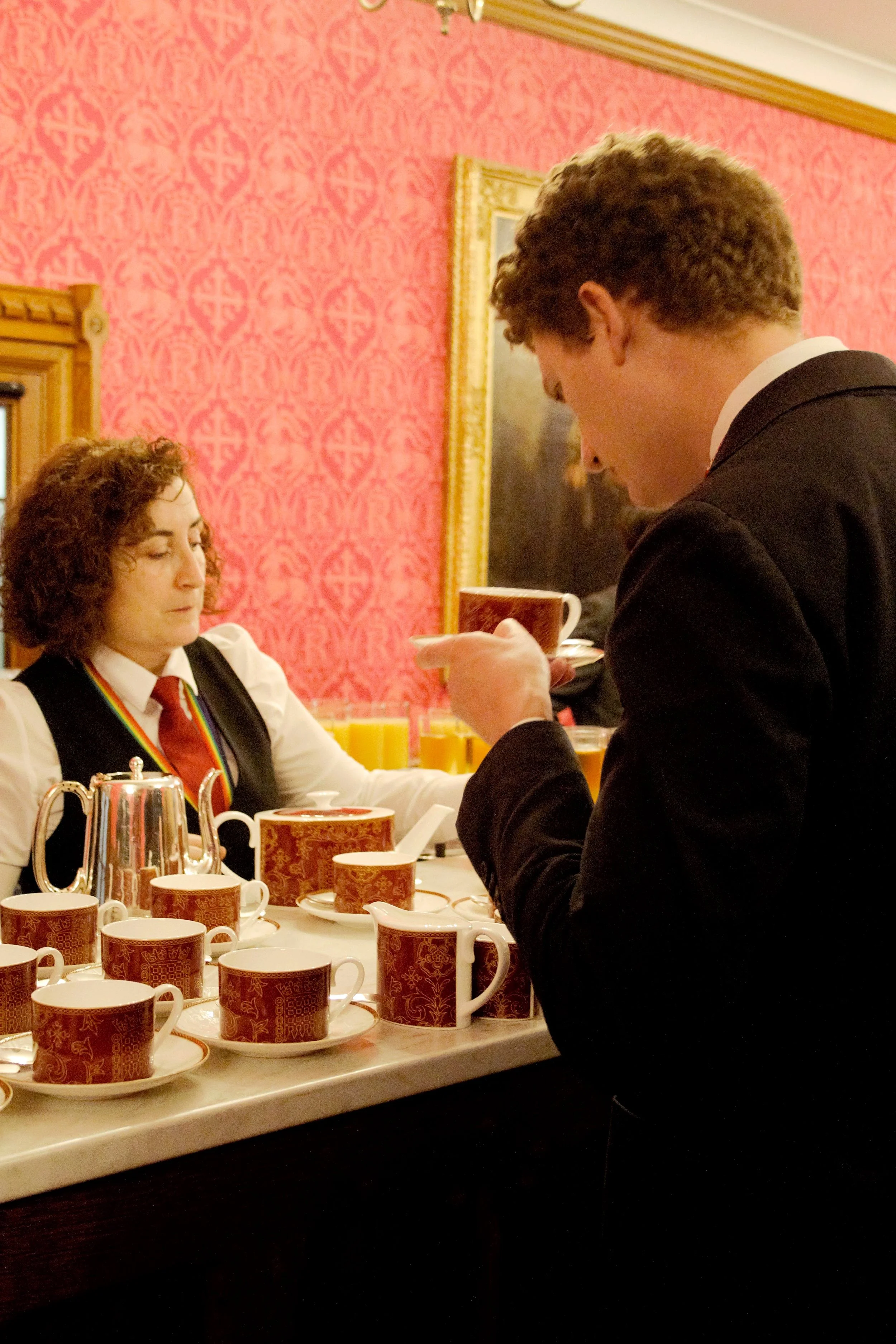 Close of shot of a guest dressed in a black suit, collecting a cup of tea and a waitress in a white shirt, black waistcoat and red tie serving the tea