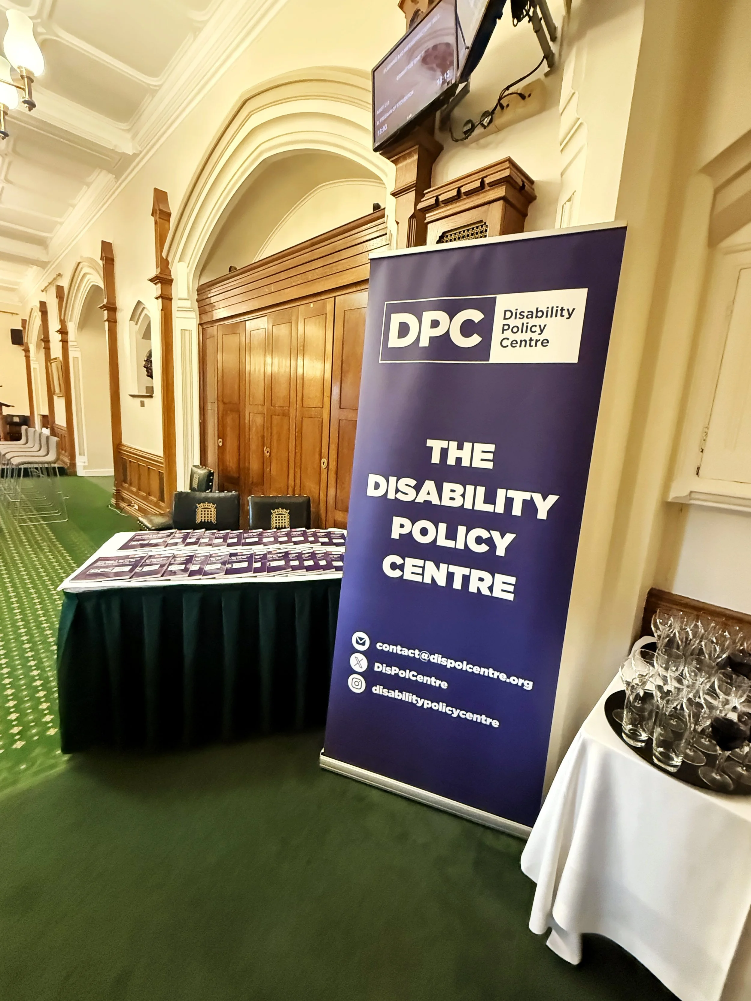 A photograph of the Churchill Room in the Houses of Parliament featuring a Disability Policy Centre banner, table with brochures, chairs, wood paneling, green carpet, and glassware on a side table beneath a TV screen.