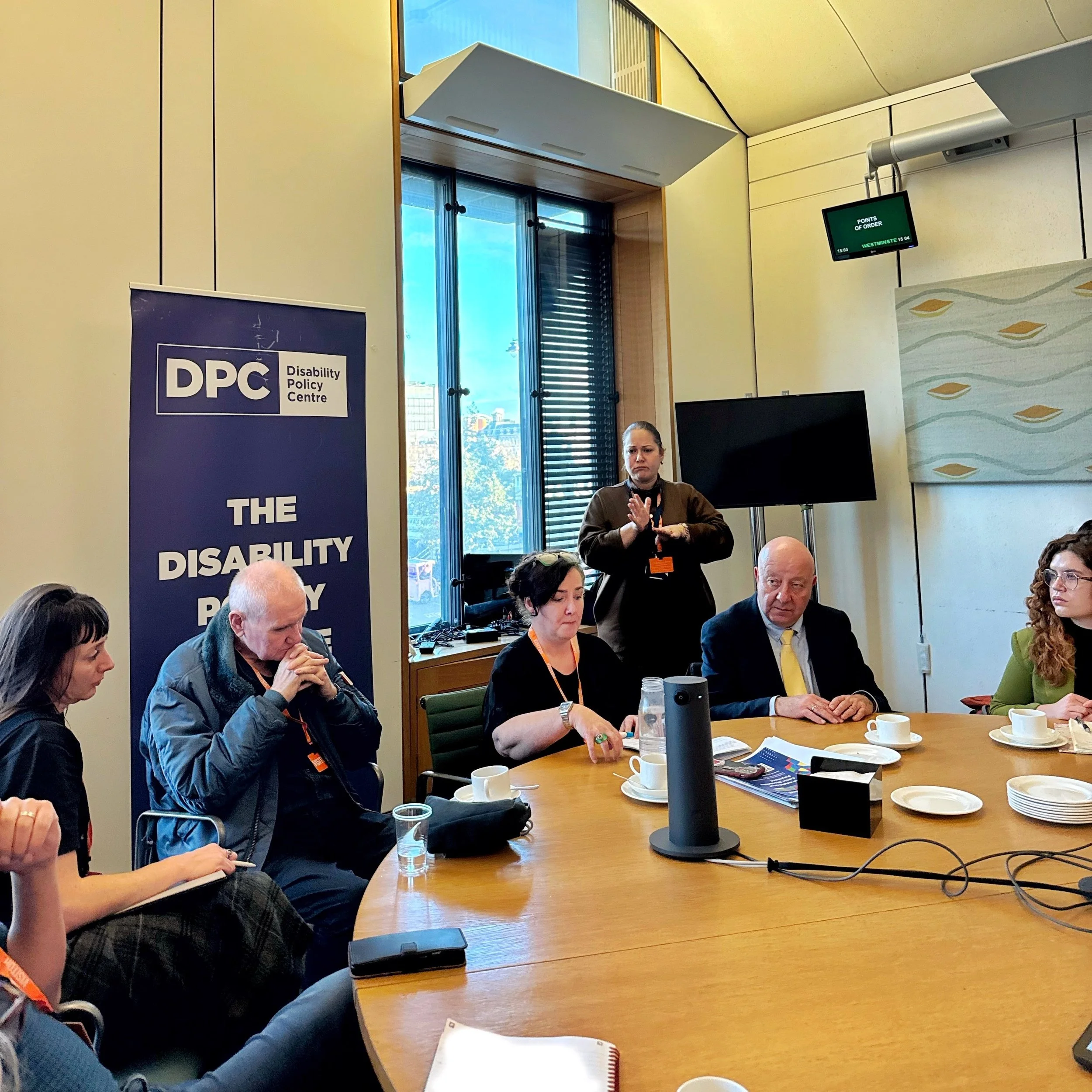 A roundtable meeting in Parliament with a DPC banner behind the group. Maria speaks on the left, Steve sits beside her in a yellow tie, Bethany in a green blazer, and Mikey in a white shirt on the right.
