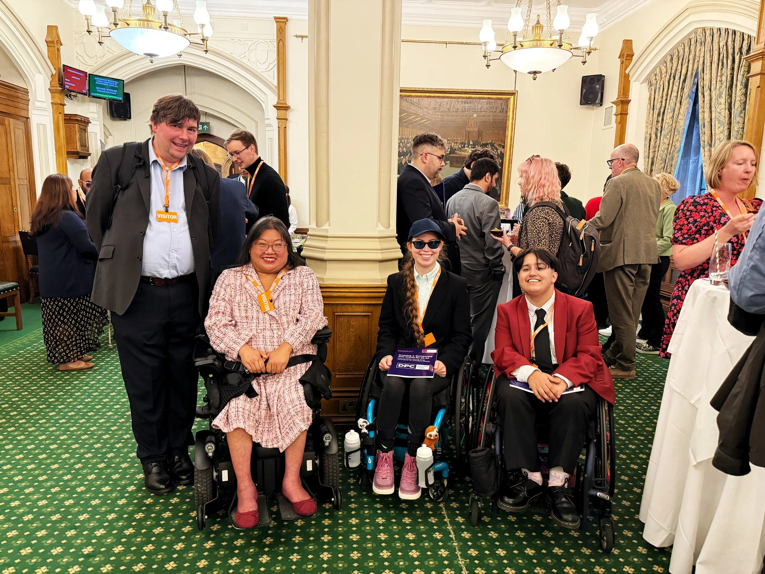 Group photo in the Churchill Room, Houses of Parliament, featuring three wheelchair users and one standing guest smiling at the camera, with other attendees networking in the background beneath chandeliers and paintings.