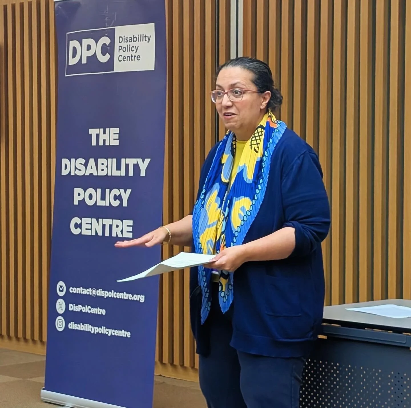 Hina stands speaking at the front of a City Hall room, wearing a blue outfit and scarf, holding papers. A Disability Policy Centre banner stands behind her during the roundtable event session.