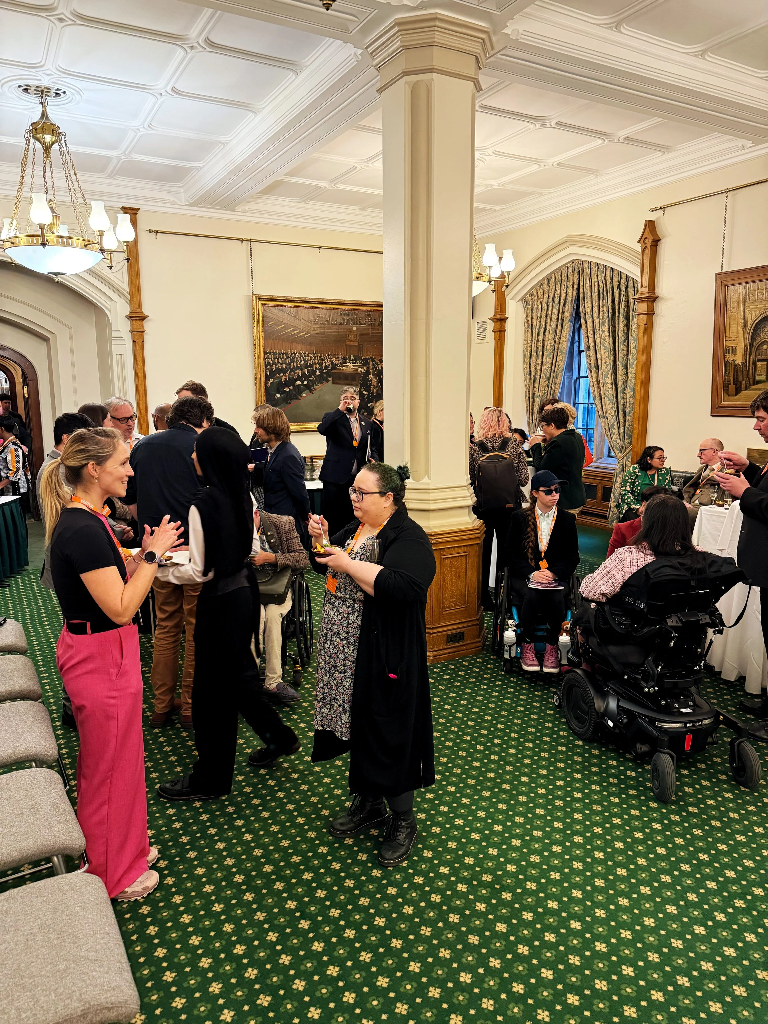 Busy scene in the Churchill Room, Houses of Parliament, with groups of people networking and talking. Several wheelchair users and seated guests are present, surrounded by chandeliers, paintings, and green patterned carpet.