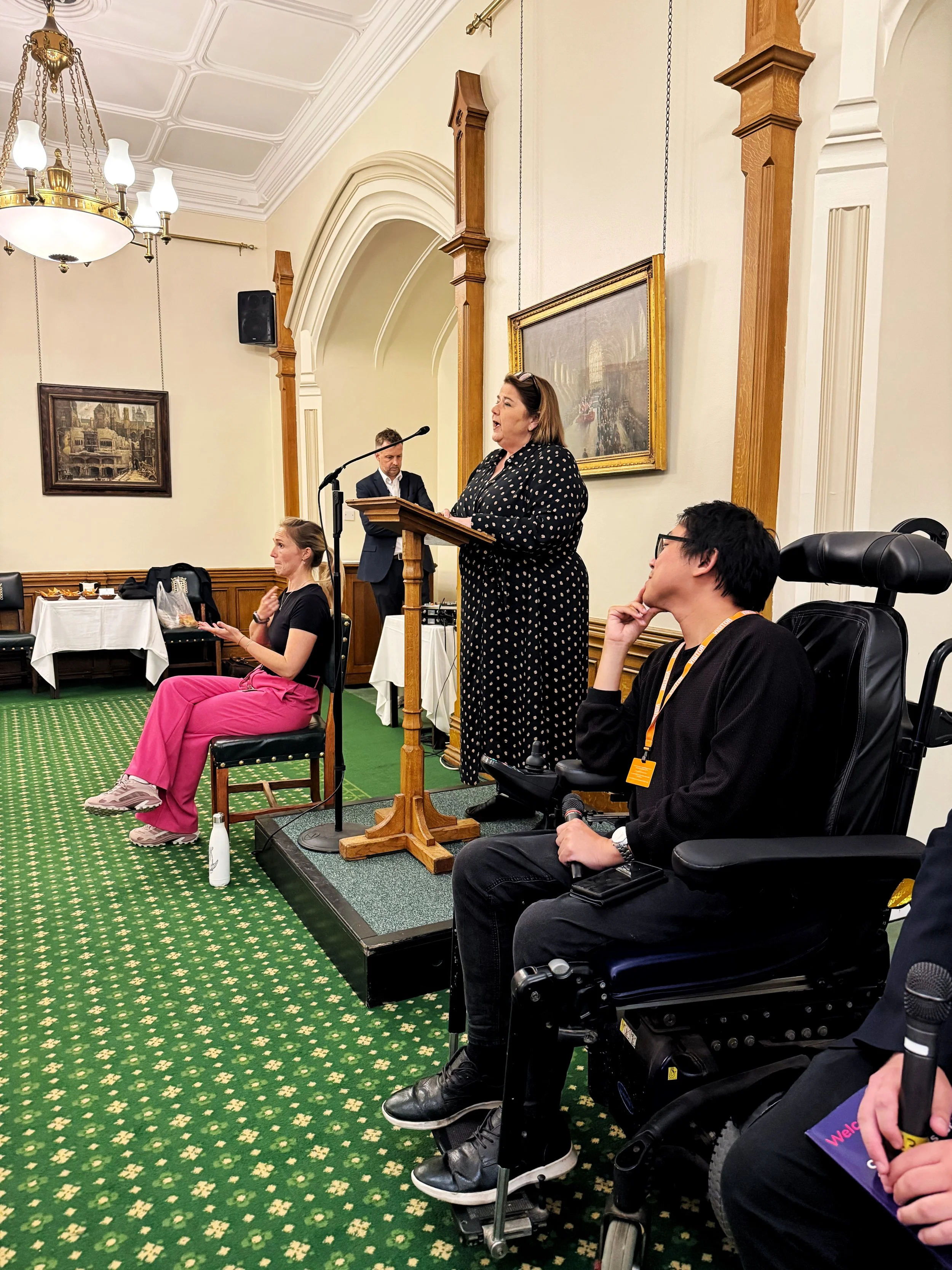 Julie Minns MP speaks at a podium in the Churchill Room, Houses of Parliament, while James Lee, the event chair, sits in a wheelchair beside her. Other panel members are seated nearby.