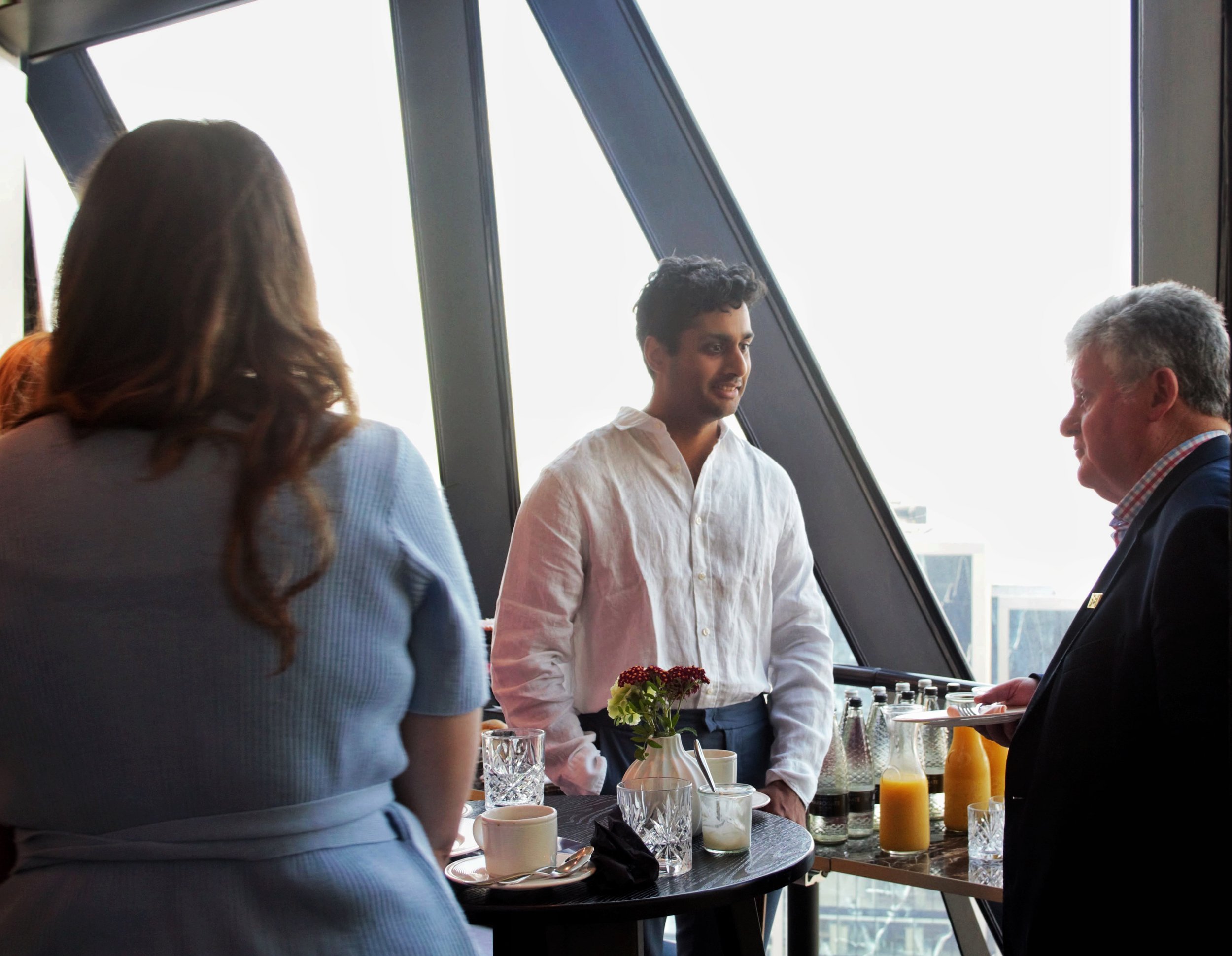 Arun and Johnny stood up and talking in front of a large glass window overlooking london.