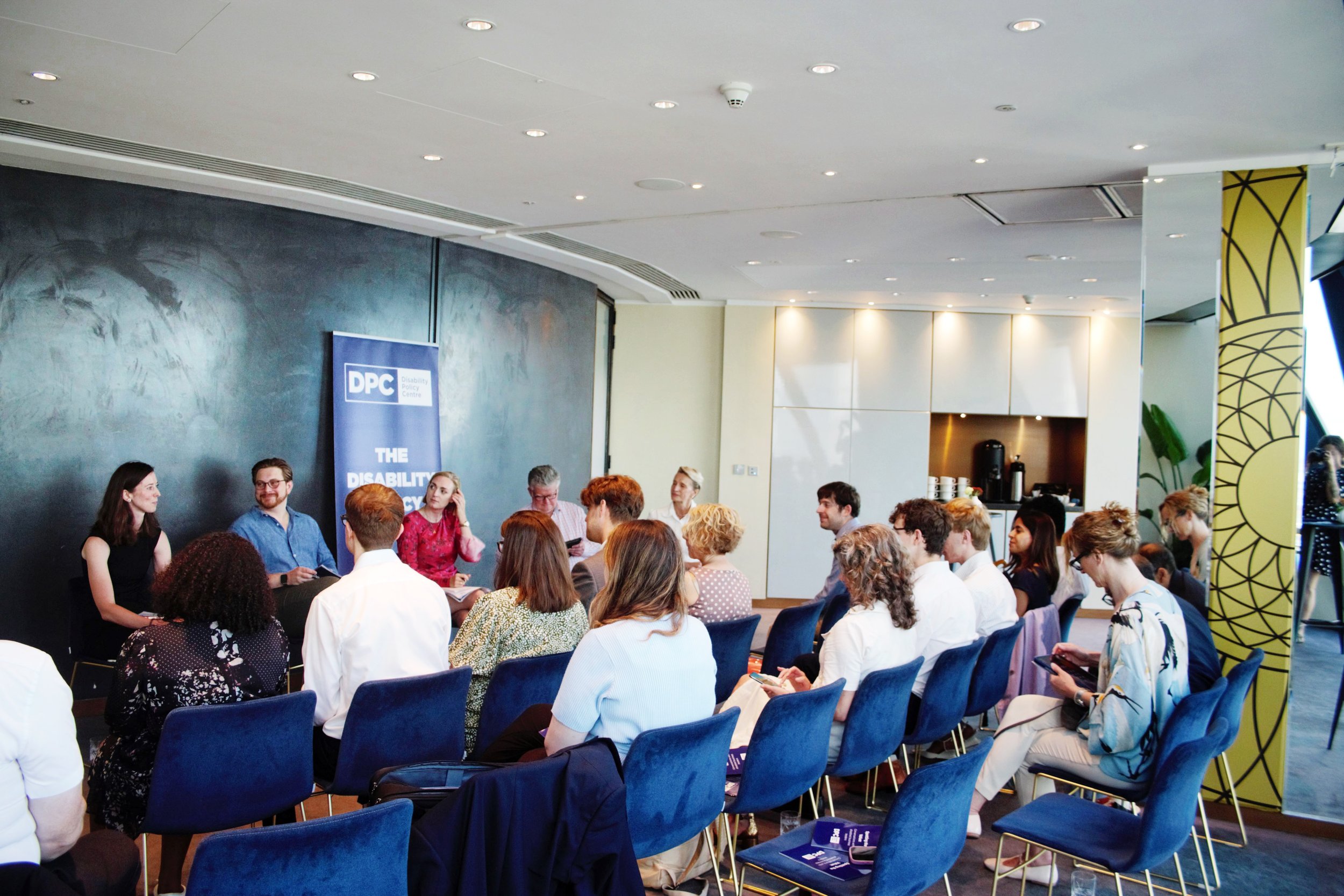 Audience members seated in rows of blue chairs attentively face a panel discussion hosted by the Disability Policy Centre, held in a modern room with ambient lighting and a casual atmosphere.