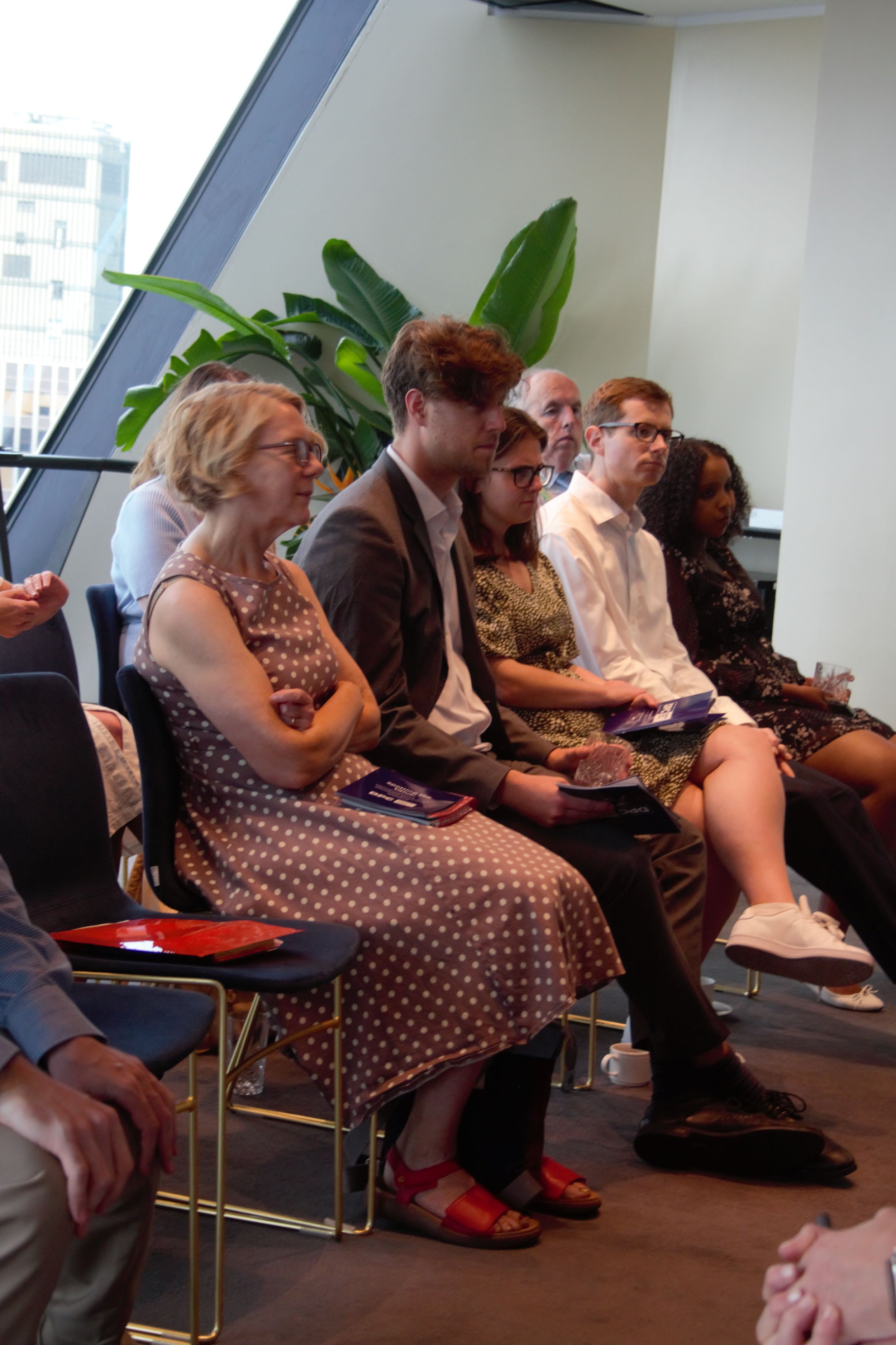 Photograph of audience sat down looking at panel. Behind is a large window and plant.