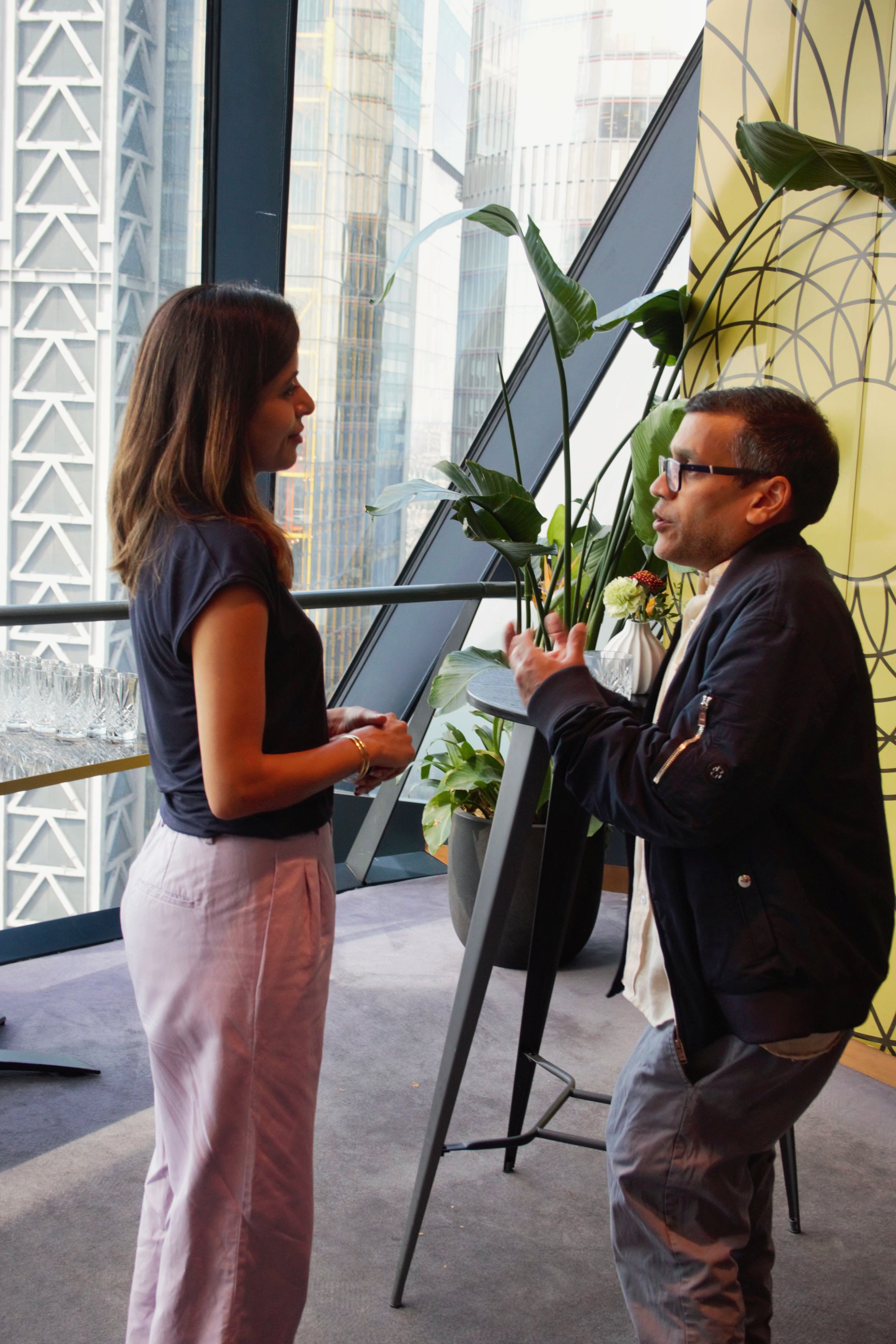 Photograph of two guest stood up, facing each other and talking in front of a large window. To the left is a female in a lilac suit and on the left is a man in a black bomber jacket and grey trousers