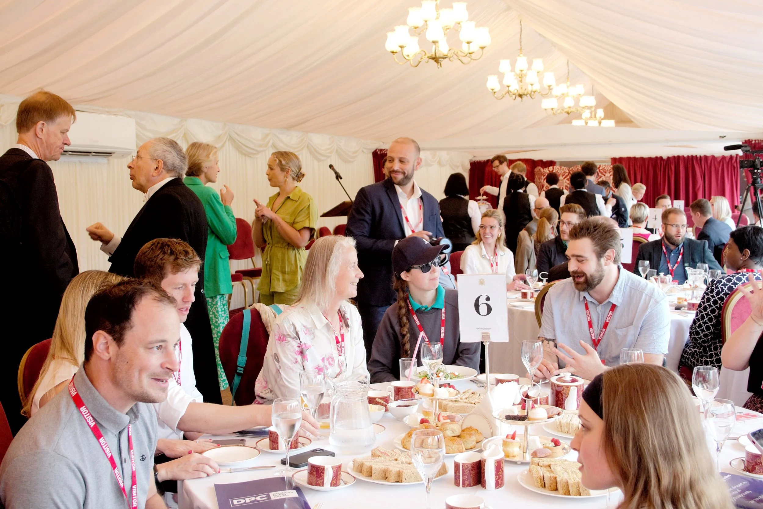 Wide shot of the entire room with half the guest sat down at their tables conversating and some stood up. Cakes and tea is served on the tables, the stage is in the background with white curtains and chandeliers on the celing