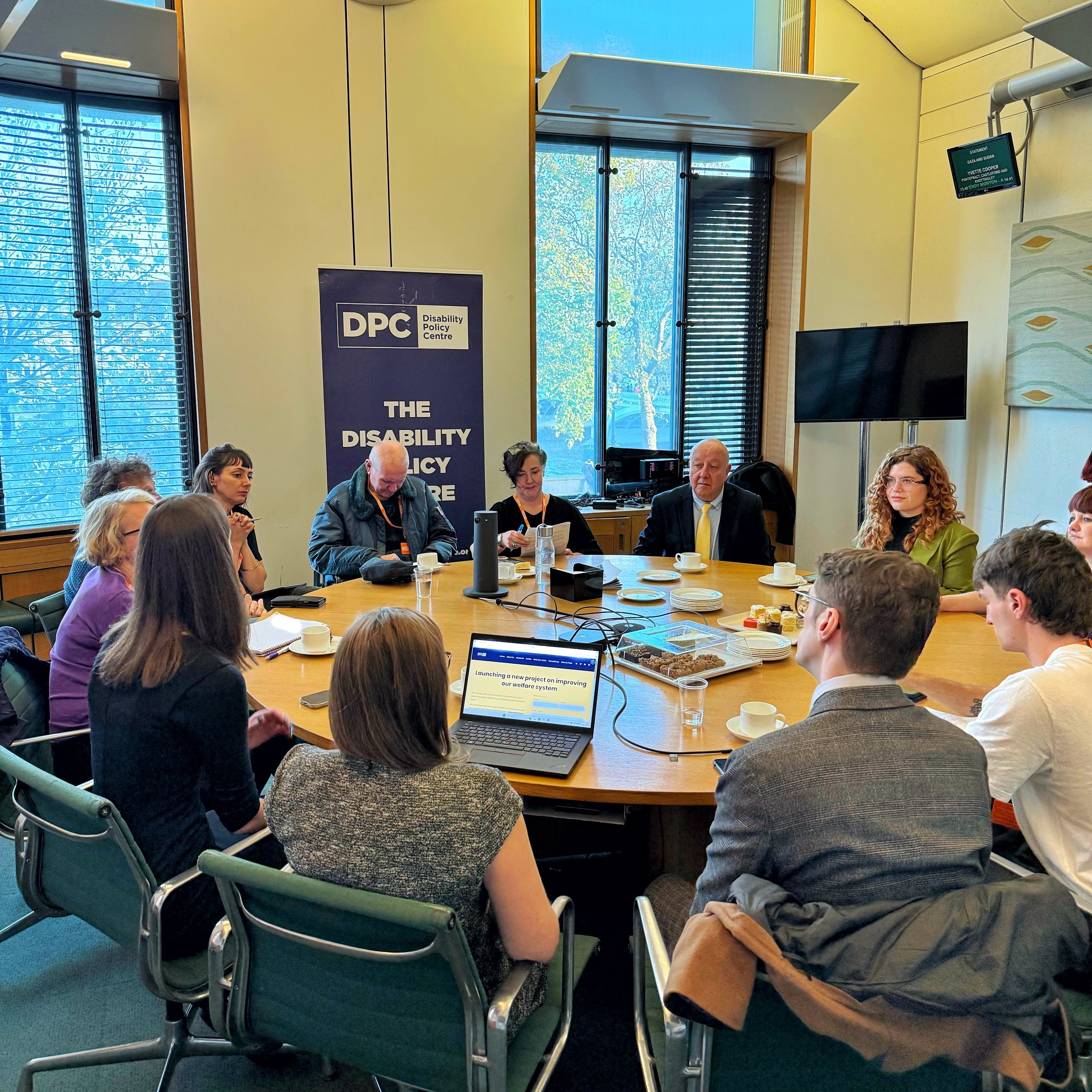 A roundtable meeting in Parliament. Chloe speaks with her back to the camera, while other speakers sit opposite her: Bethany on the back right, with Steve and Maria seated to her left. Attendees listen around the table.