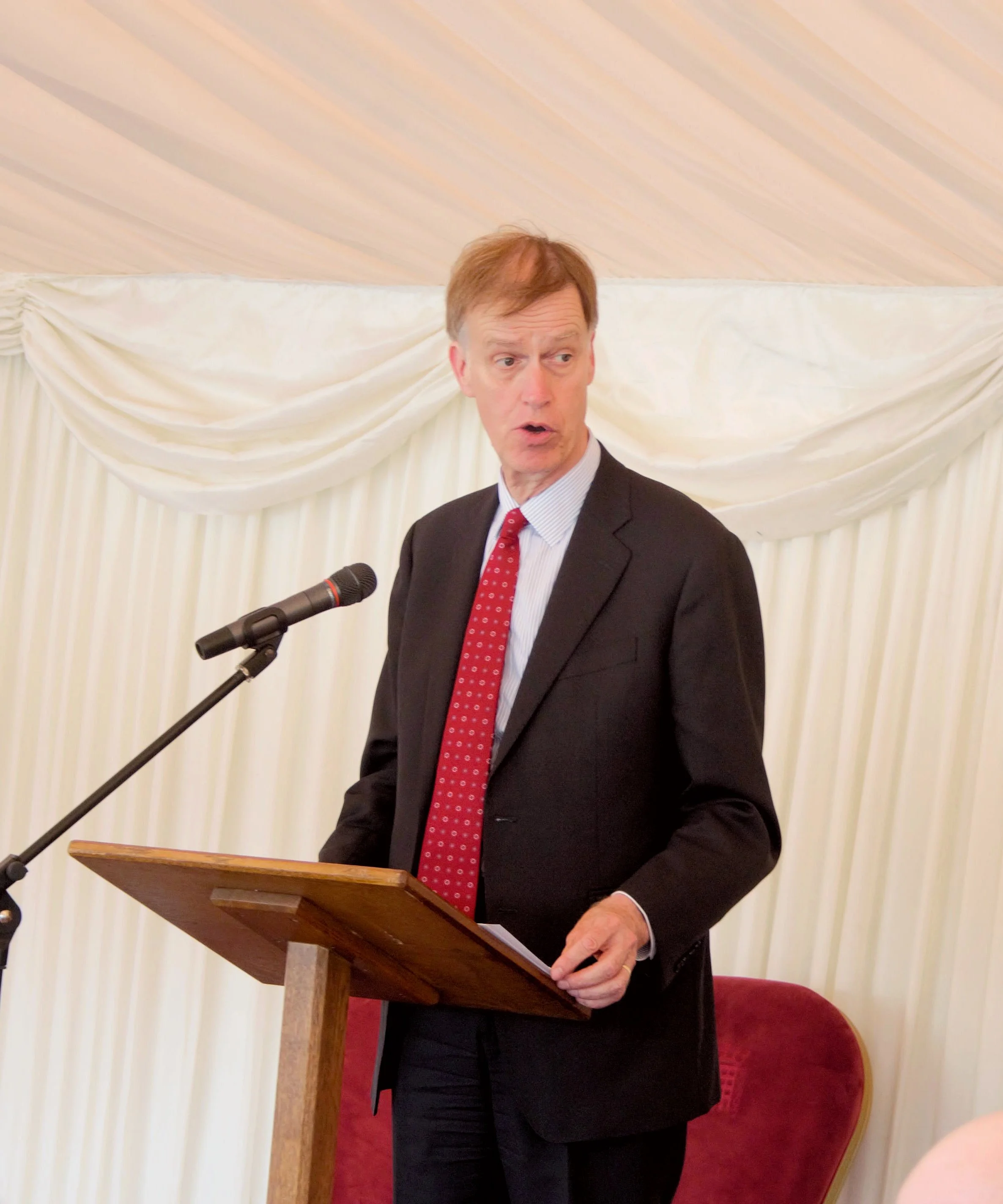 Close up shot of Sir Stephen Timms MP speaking on the stage infront a podium and microphone, he wearing a black suit with a white shirt and red tie