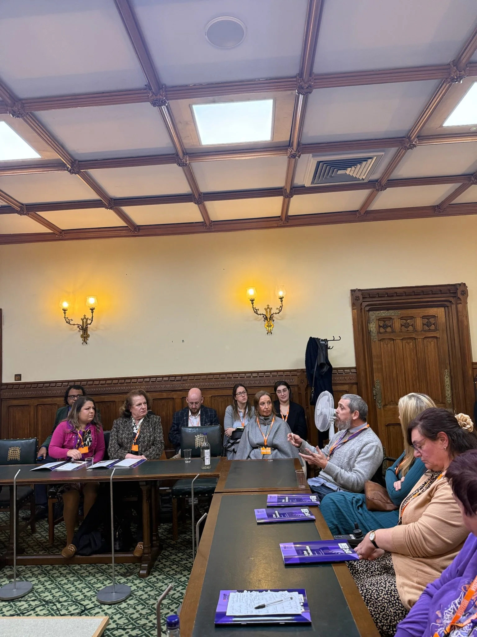 An attendee speaks during the discussion while others listen around the table, with A Street Transformation reports and notes visible on the desks in the parliamentary room.