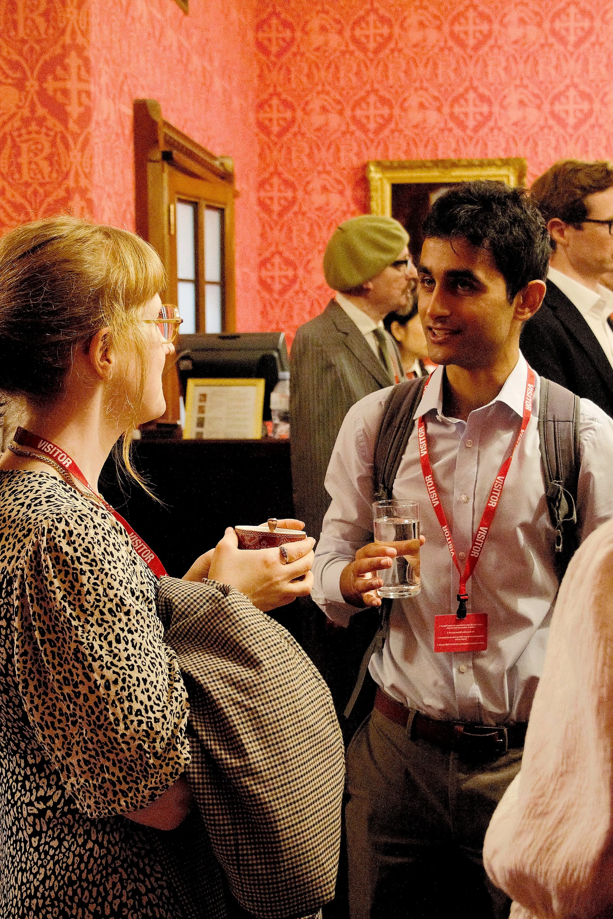 Close up of two guests speaking in the red room. the guest to the right is a male with black hair, wearing a ligh tblue shirt with dark grey trousers, a backpack and a red visitos langyard. the guest to the left is a female turned to her side wearing