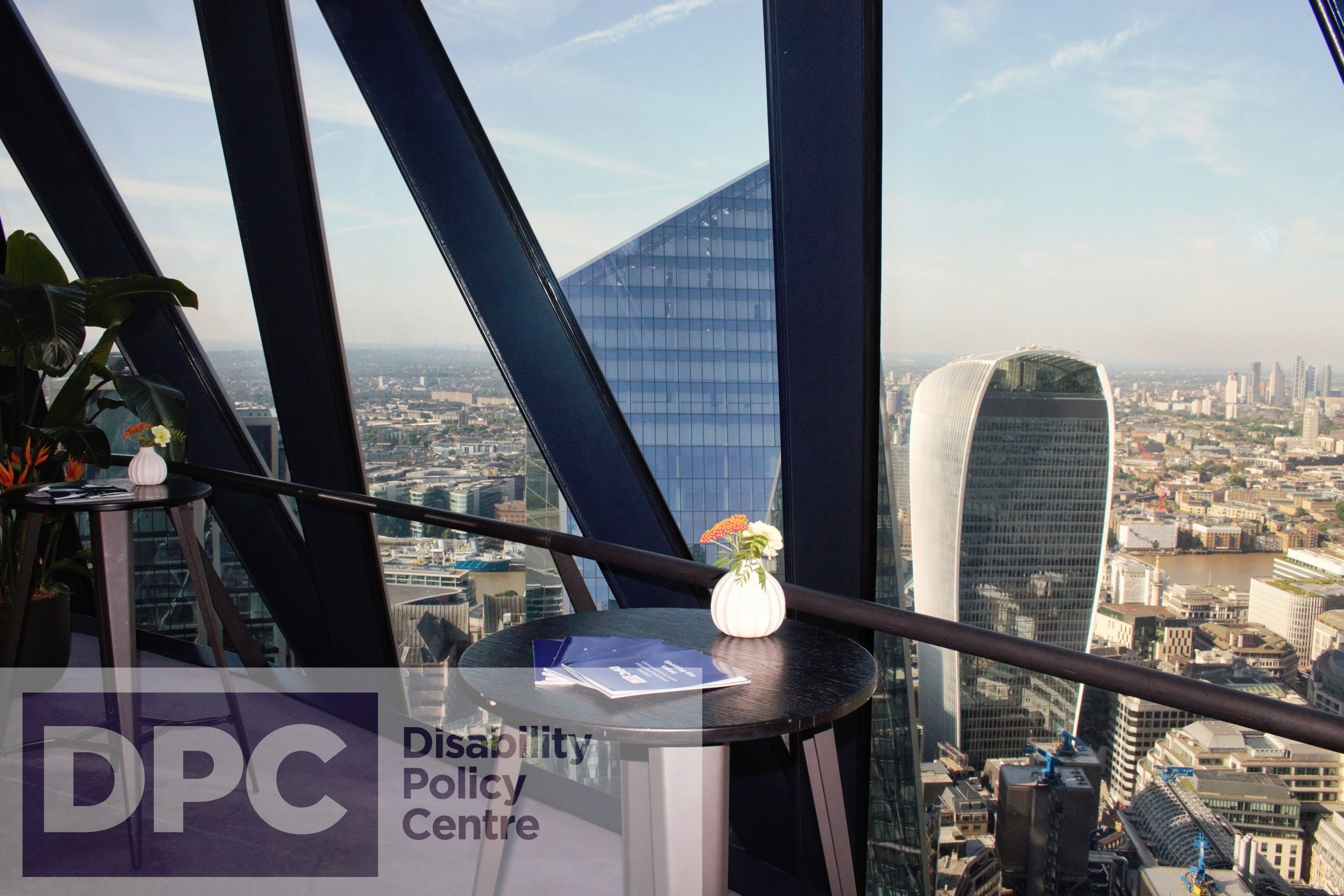 A small round table with Disability Policy Centre booklets and a flower vase overlooks London’s skyline through large windows, featuring iconic buildings like the Walkie Talkie and angular skyscrapers.
