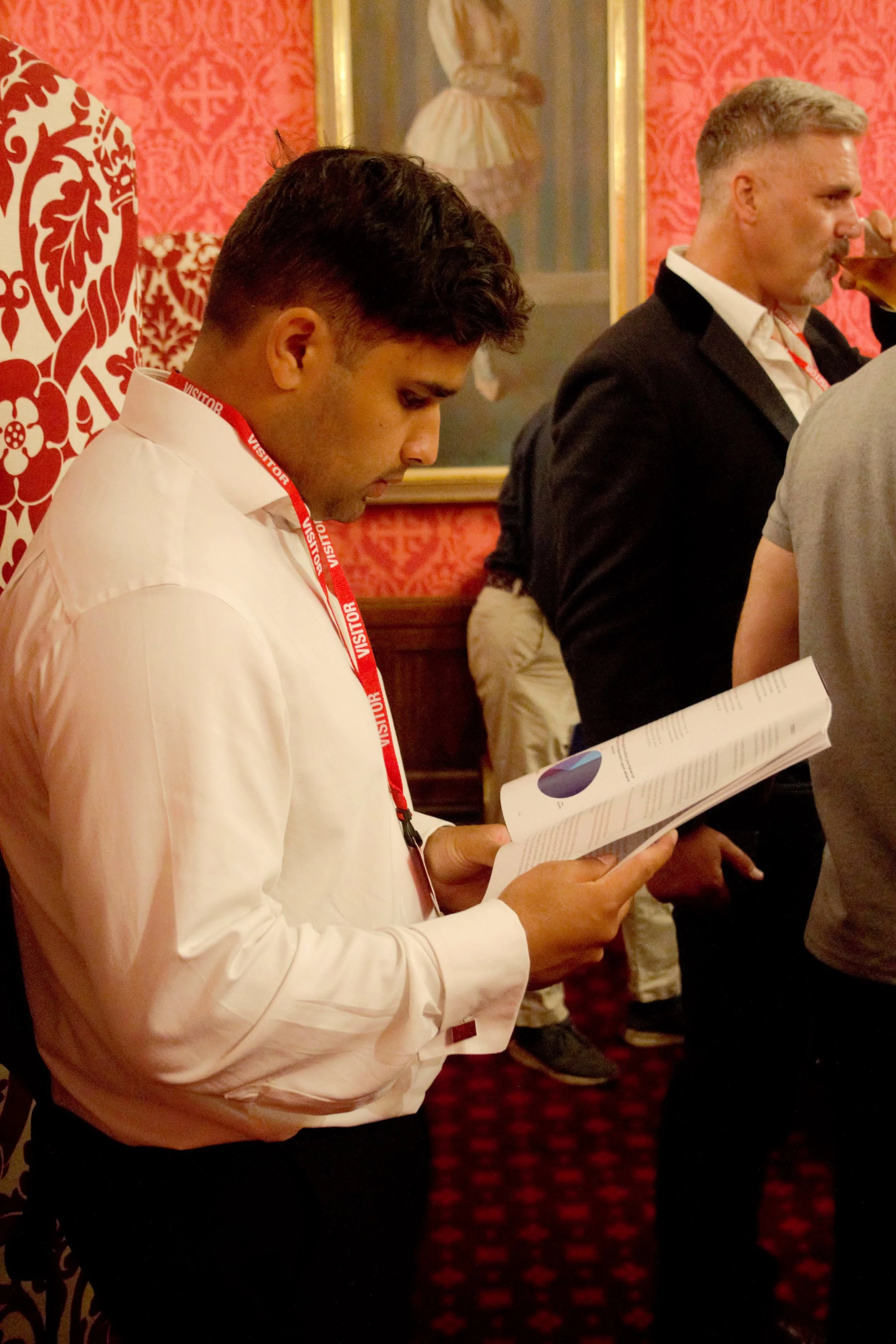 Close up shot of a guest reading the printed power of sport report in the red room. He is dressed in a white shirt and black trousers with a red visitors langyard. There are other guests in the background