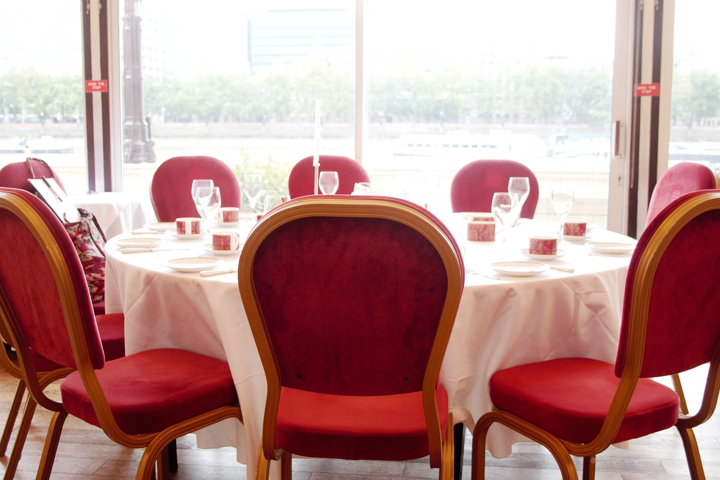 Photograph of and empty round table, that has been set up, with red velvet chairs around it and glasses set, the table is facing the window overlooking the river