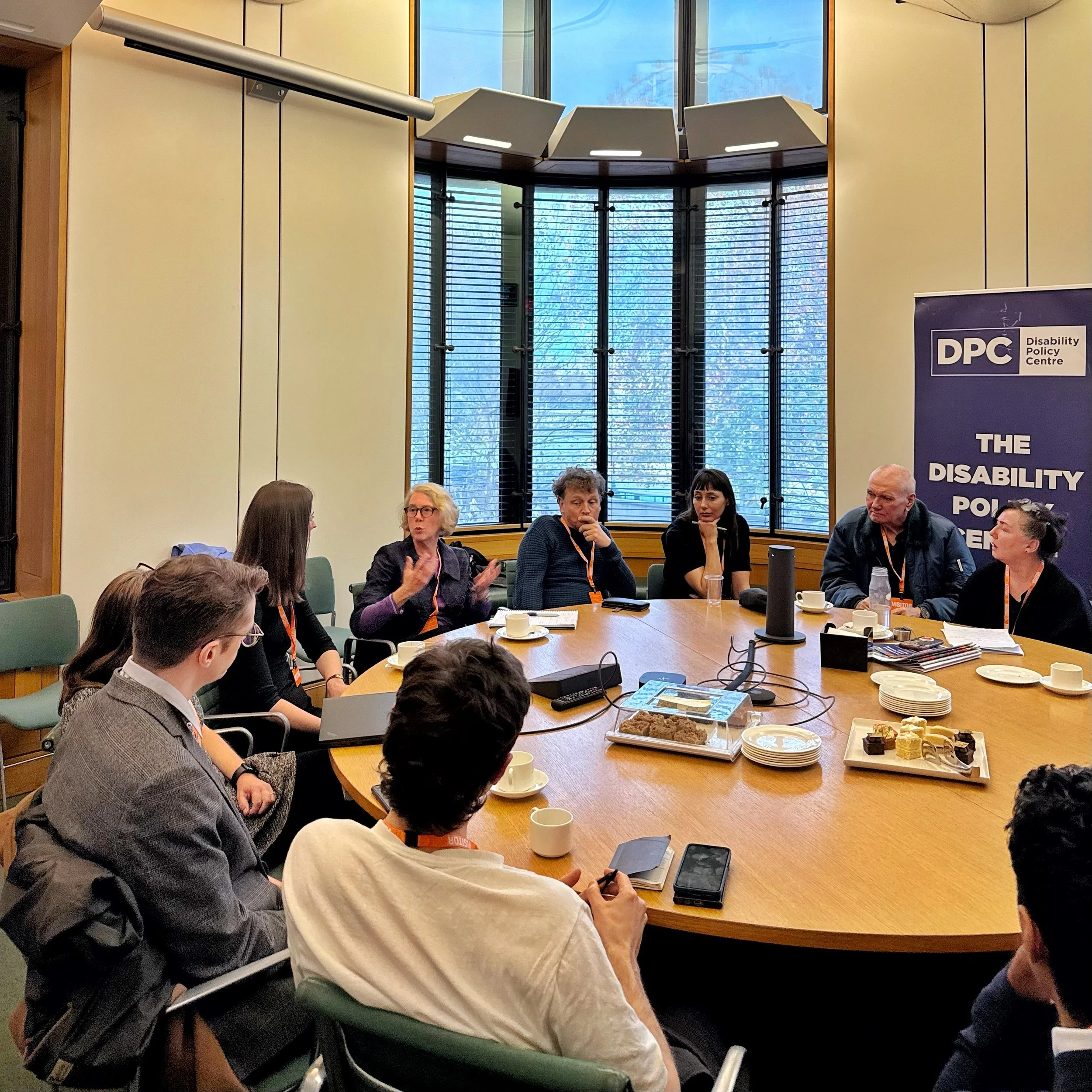 A roundtable meeting in Parliament. Joanna is speaking to the group while attendees listen around the table. The DPC banner stands on the right, with notes, drinks and refreshments spread across the table.