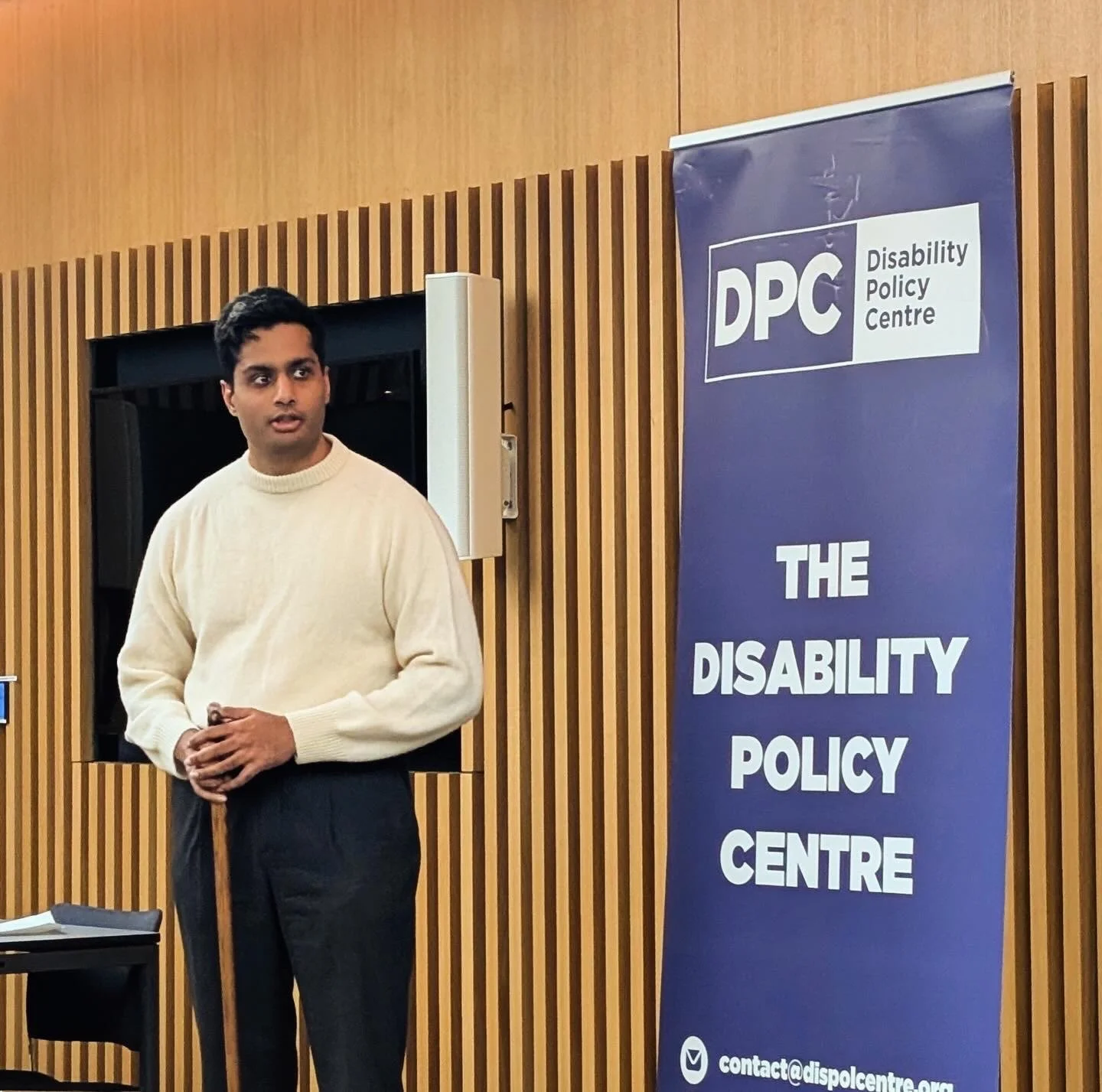 Arun stands beside a Disability Policy Centre banner at City Hall, wearing a white jumper, addressing attendees during a roundtable discussion in a modern meeting room with participants seated nearby.