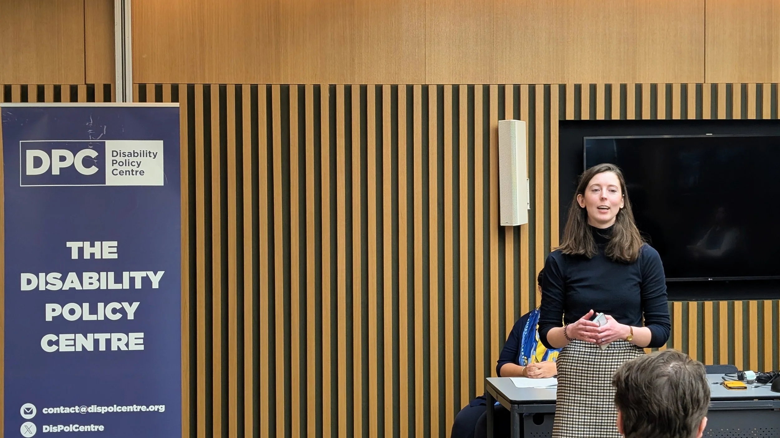 Chloe stands speaking at the front of a City Hall room, wearing a black turtle next jumper and checkered skirt. A Disability Policy Centre banner stands behind her during the roundtable event session.
