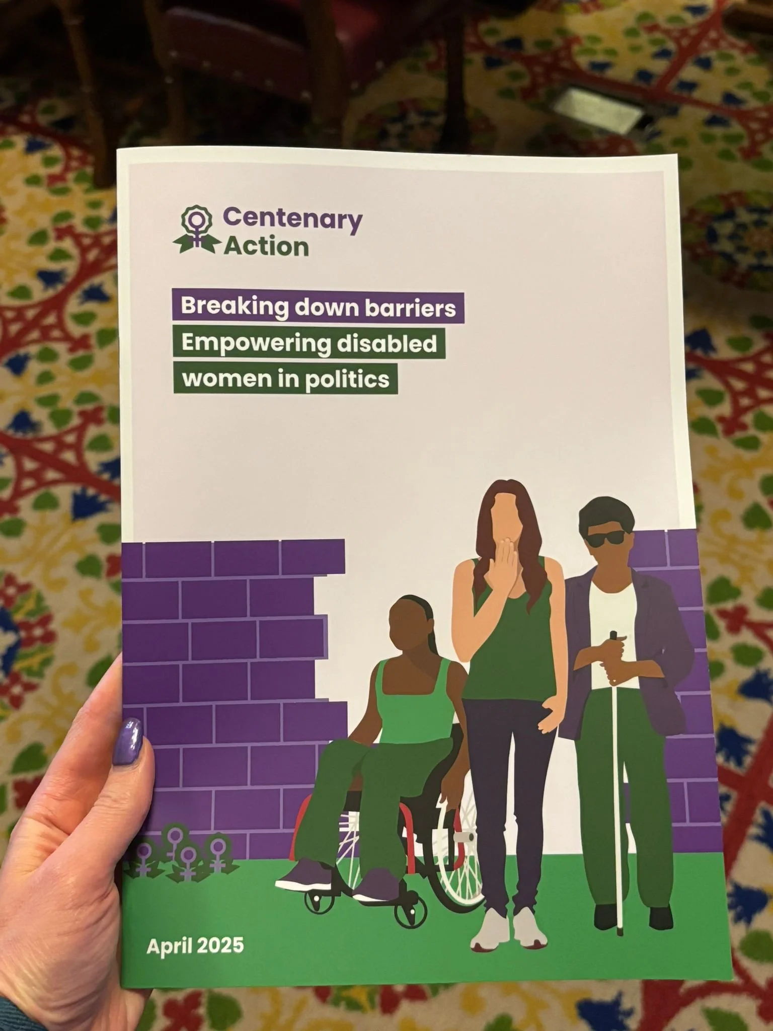 A hand holds a report titled “Breaking down barriers: Empowering disabled women in politics” by Centenary Action. The cover shows three diverse disabled women before a broken purple brick wall, dated April 2025.