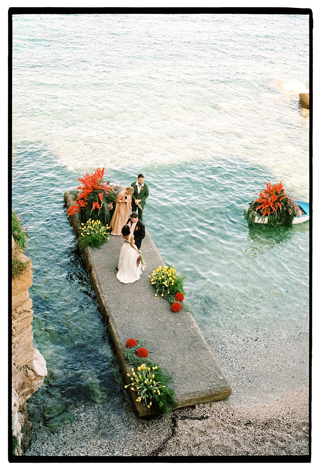 A wedding ceremony taking place on a concrete pier extending over the water, decorated with bright red, yellow, and orange flowers, with a boat nearby also decorated with flowers.