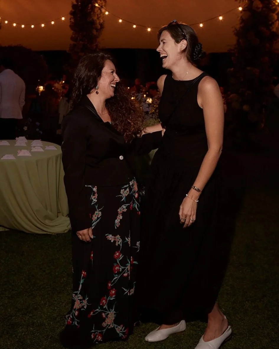 Two women laughing and talking at an outdoor evening event with string lights overhead.