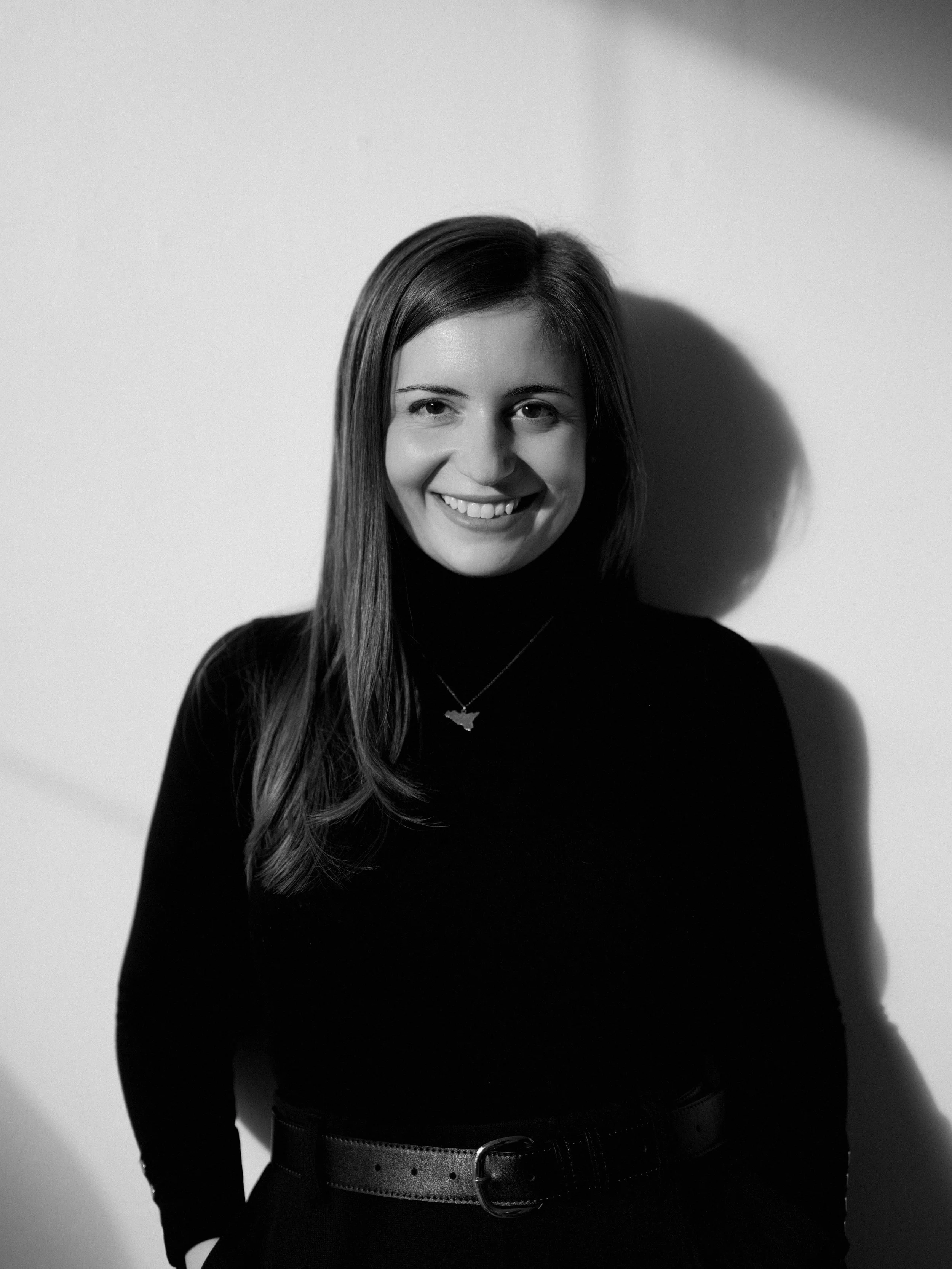 Black and white photo of a smiling woman with long hair, wearing a black turtleneck and a necklace with a fish-shaped pendant, standing against a plain wall with her shadow cast to her right.