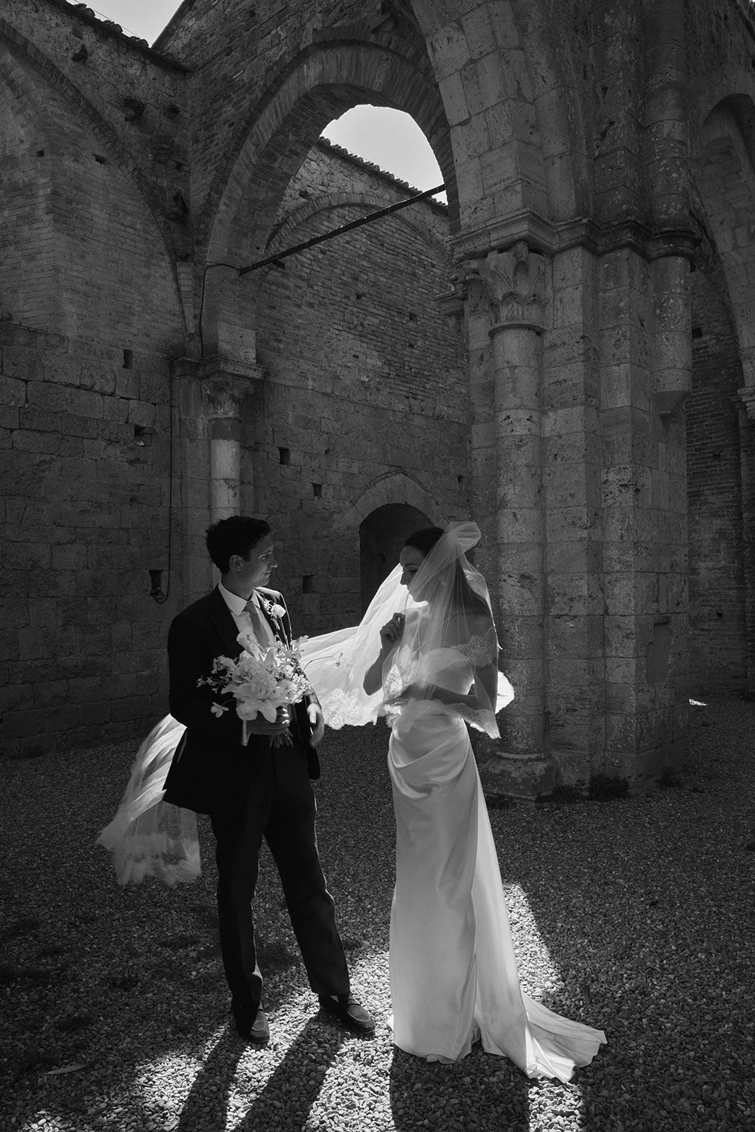 A black and white photo of a bride and groom standing inside a historic stone building. The groom holds a bouquet of flowers, and the bride wears a long dress and veil, with sunlight illuminating them from above.