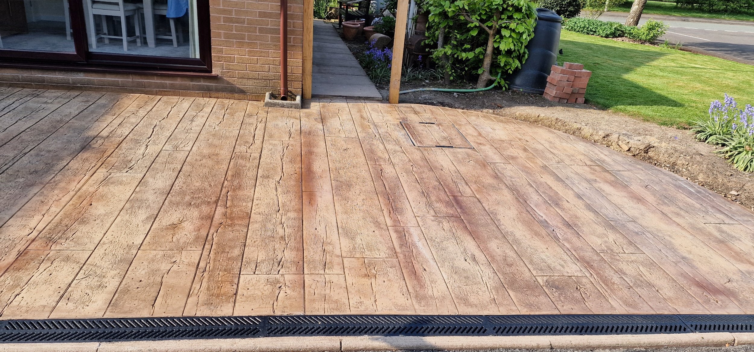 Reclaimed Timber printed concrete in biscuit. Linear drains leading to soak away make this Project SUDS compliant and a later addition of a low wall rendered in wallcrete and printed on all aspects to resemble timber and a lich gate were added