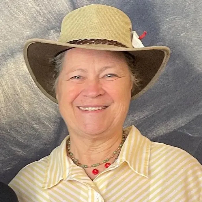 Woman wearing a tan hat and striped shirt, smiling against a gray background.