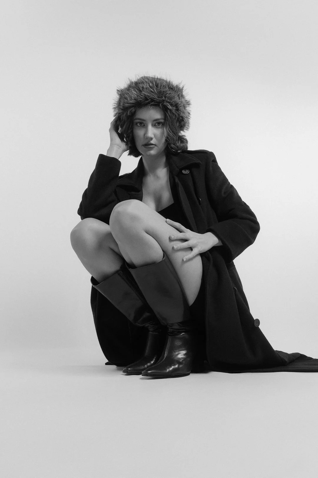 Une femme pose dans une tenue élégante, portant un manteau noir, des bottes en cuir et un bonnet en fourrure, dans une photo en noir et blanc.